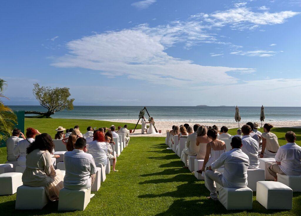 Couple exchanging vows during a wedding ceremony on the lush lawn of Casa Escondida, with the Pacific Ocean beach as a stunning backdrop in Punta Mita.