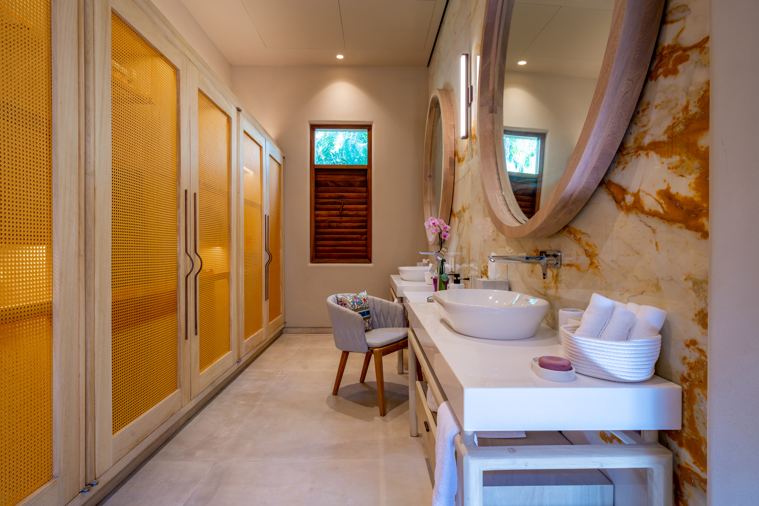 Narrow ensuite bathroom showing a double-vanity with vessel sinks on a white counter, amber-lit cane wardrobe doors along the opposite wall, a round marble-framed mirror, and a small louvered window bringing in garden light, Casa Akama, Ranchos Estates, Punta Mita.