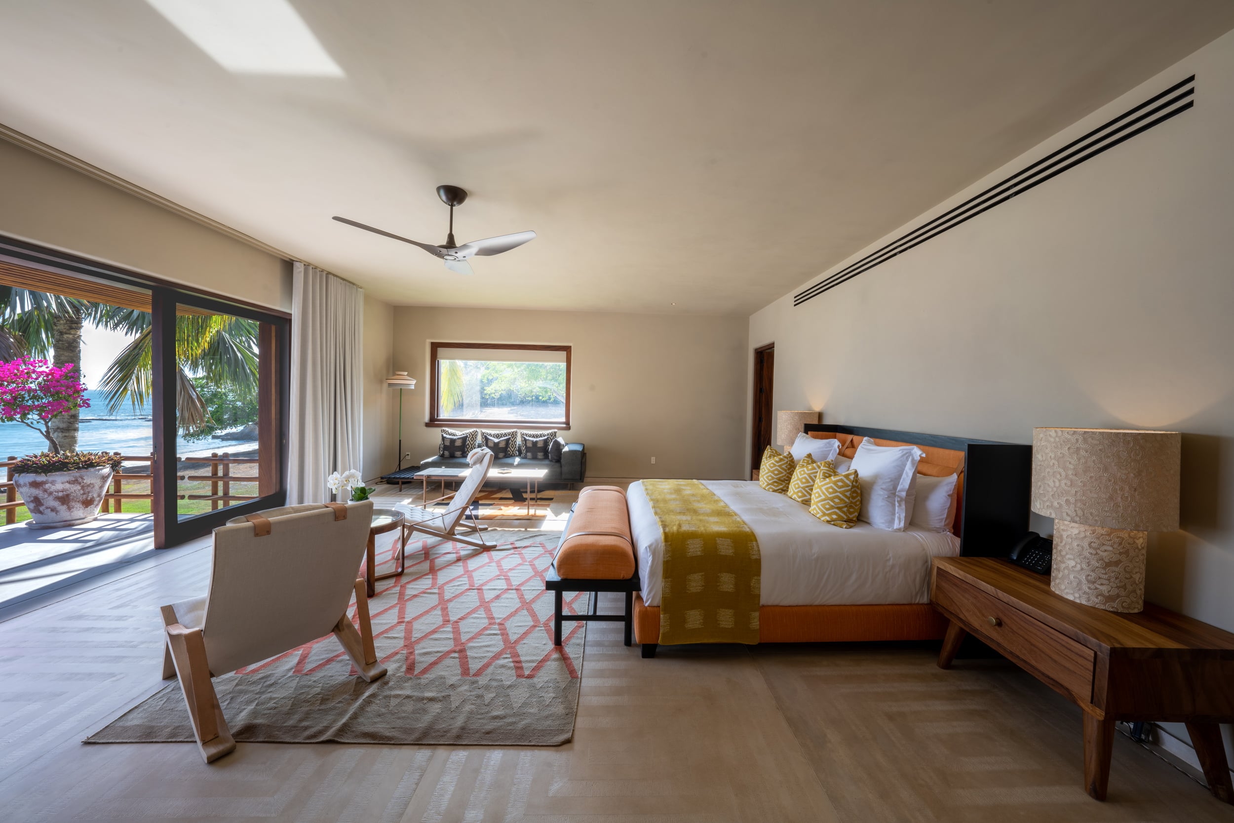 King bedroom with ocean view through full-height sliding glass doors opening to a terrace framed by palm trees and bougainvillea, furnished with canvas sling chairs, an orange upholstered bench, and a woven pendant lamp on the nightstand, Casa Akama, Ranchos Estates, Punta Mita.