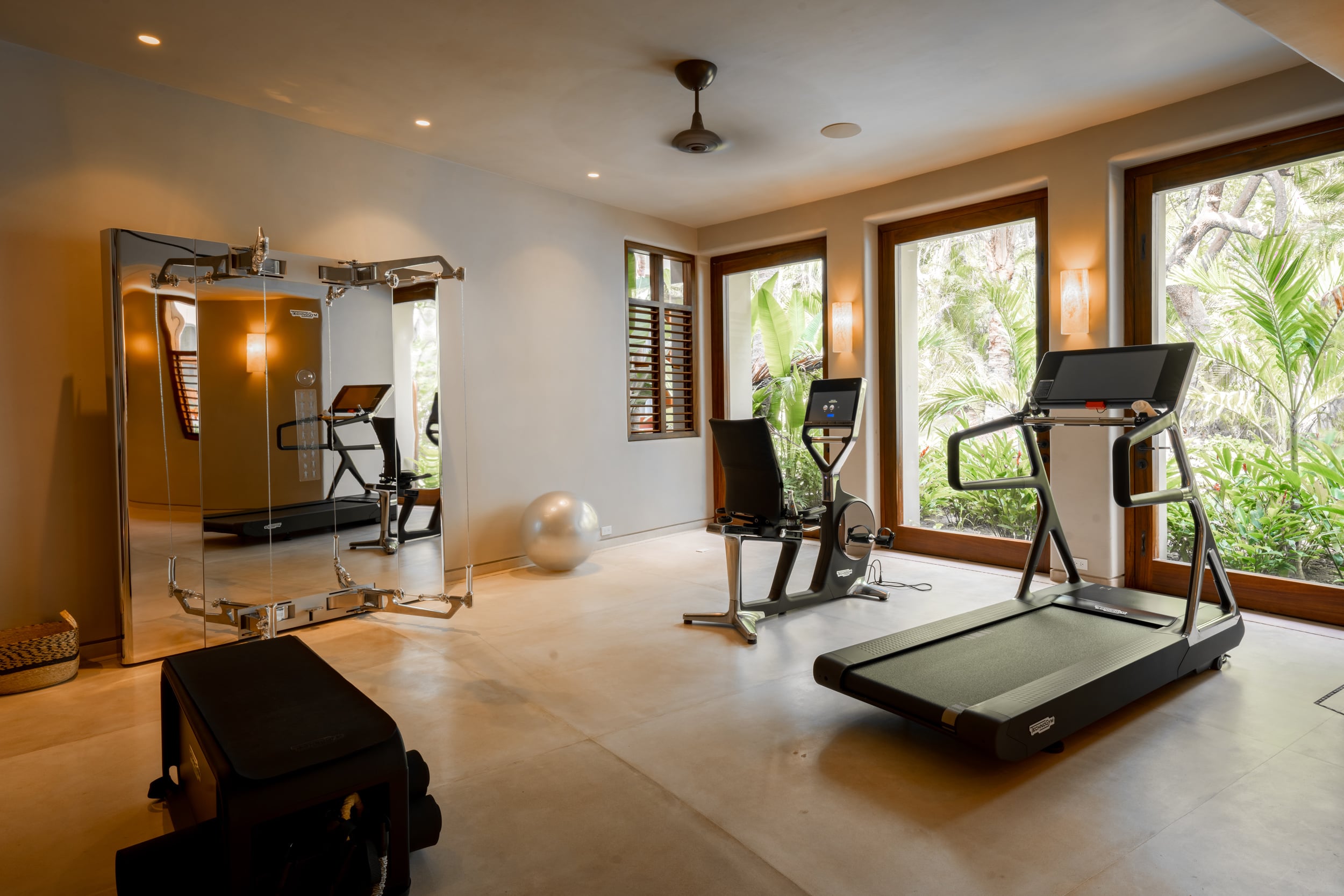 Private fitness room with a treadmill and stationary bike positioned in front of large louvered windows overlooking tropical garden, a floor-to-ceiling mirror on the opposite wall reflecting cable-machine equipment, Casa Akama, Ranchos Estates, Punta Mita.