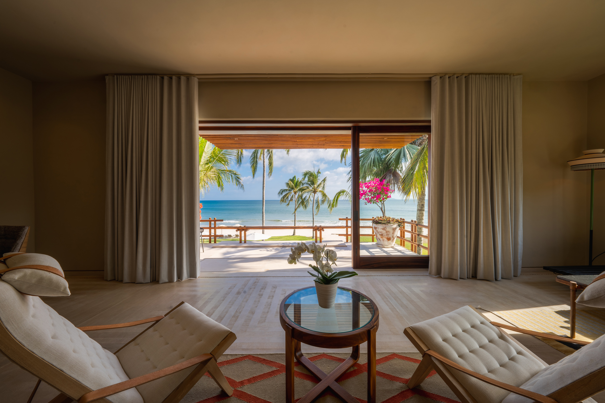 Interior sitting area with two tufted white armchairs and a round glass coffee table centered on a patterned rug, framed by floor-to-ceiling white linen drapes drawn back to reveal a wide terrace, palm grove, and the Pacific Ocean beyond, Casa Akama, Ranchos Estates, Punta Mita.