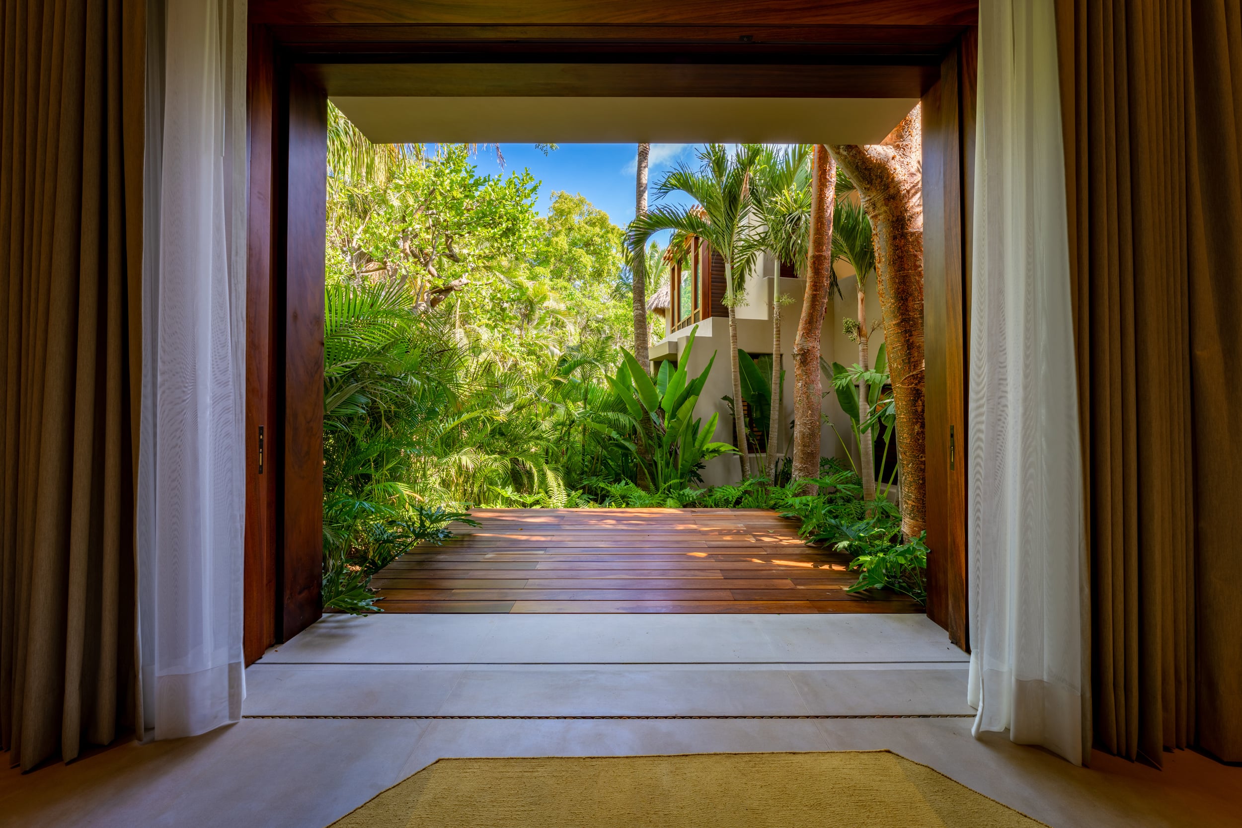 View from inside a room through a large square opening framed in dark wood, looking out along a hardwood-deck path bordered by dense tropical planting of palms and broad-leaf foliage toward the villa structure beyond, Casa Akama, Ranchos Estates, Punta Mita.