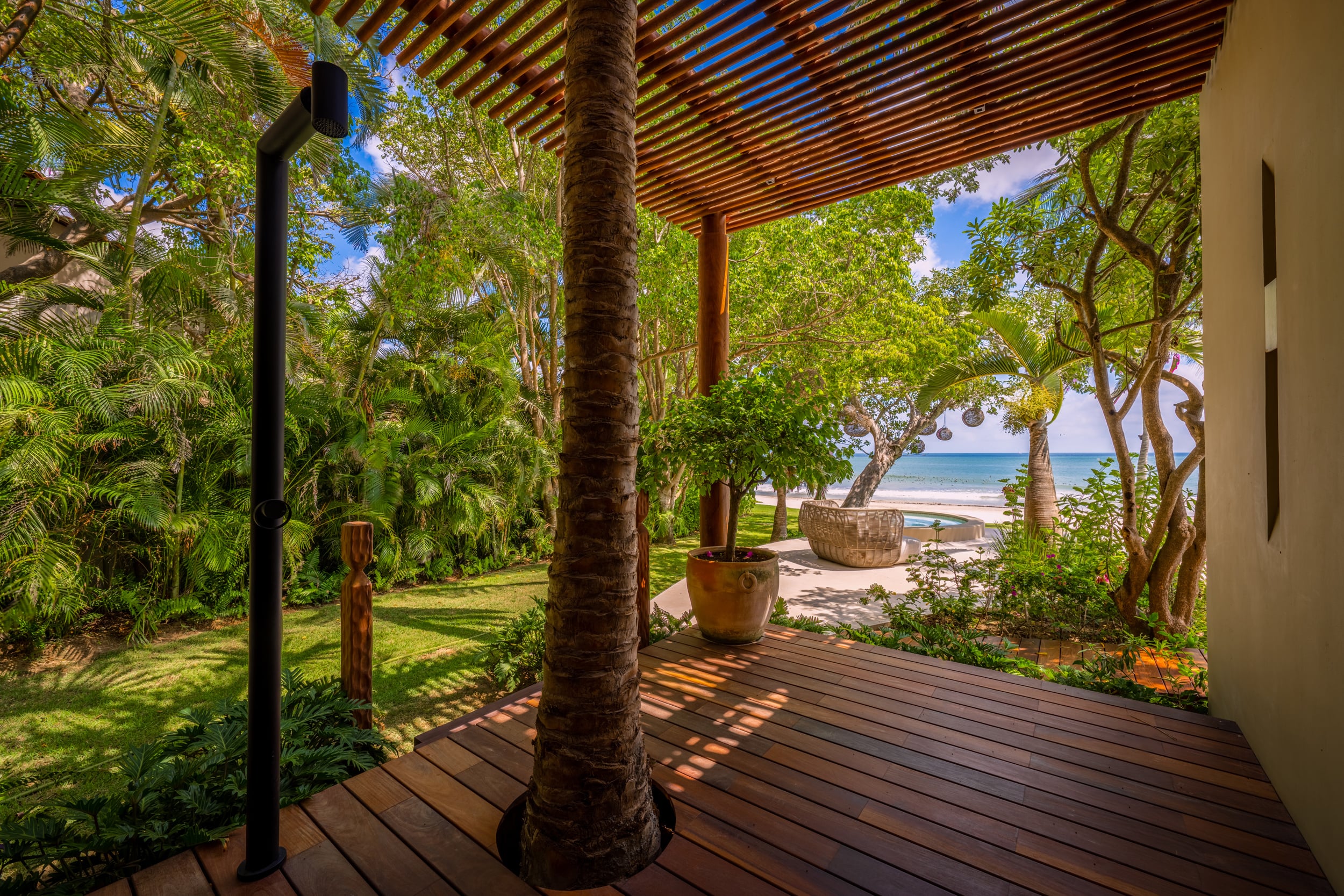 Covered hardwood deck with a pergola-style slatted roof, a palm trunk growing through the decking, a terracotta planter, and a view through the foliage to the beach and Pacific Ocean with a woven-reed chair visible at the shore, Casa Akama, Ranchos Estates, Punta Mita.