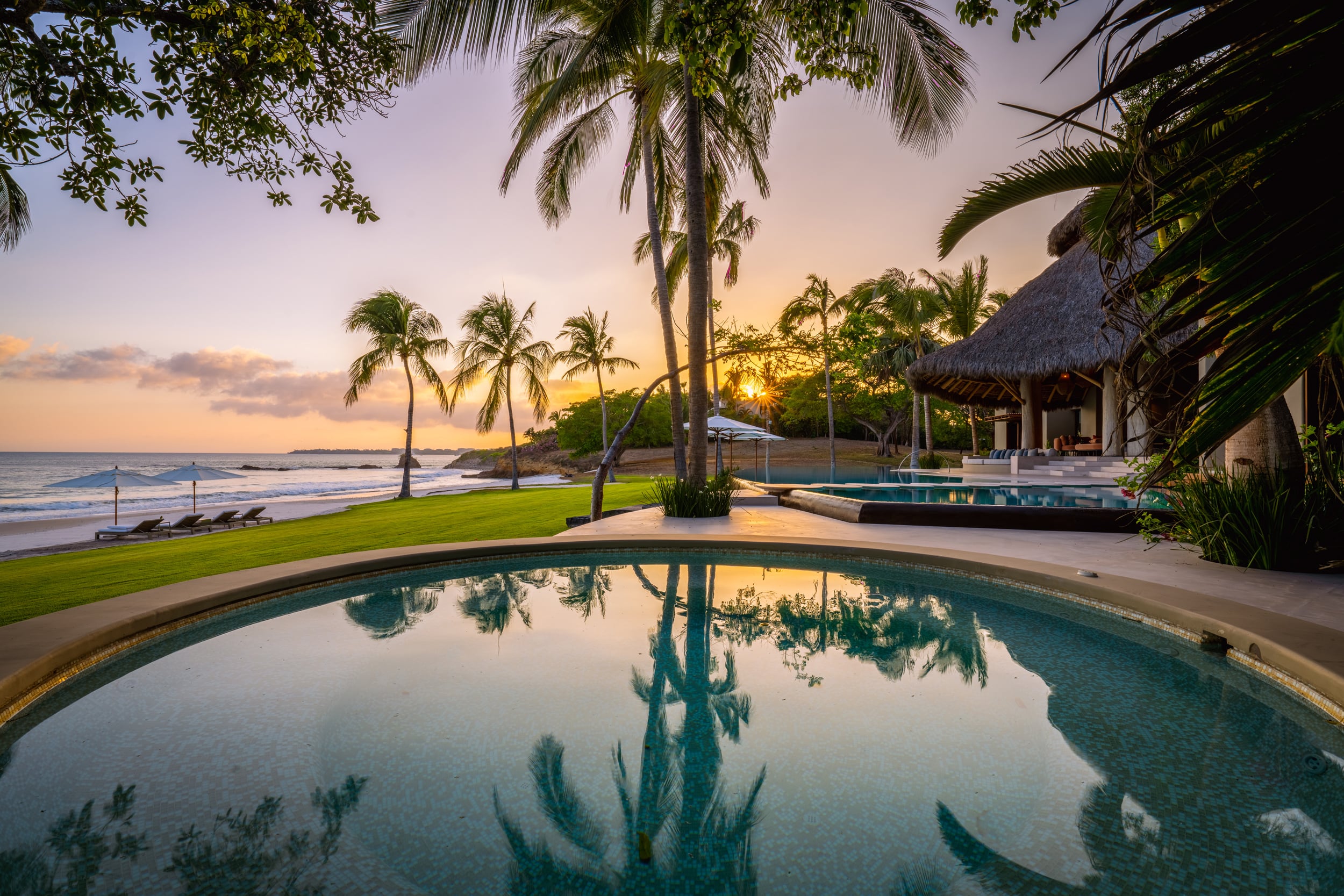 Sunset view from the pool area showing calm water reflecting palm silhouettes and an orange-and-purple sky, a palapa-roofed pavilion to the right, and a shallow wading pool or spa adjacent to the main infinity pool, with lounge chairs visible on the lawn, Casa Akama, Ranchos Estates, Punta Mita.