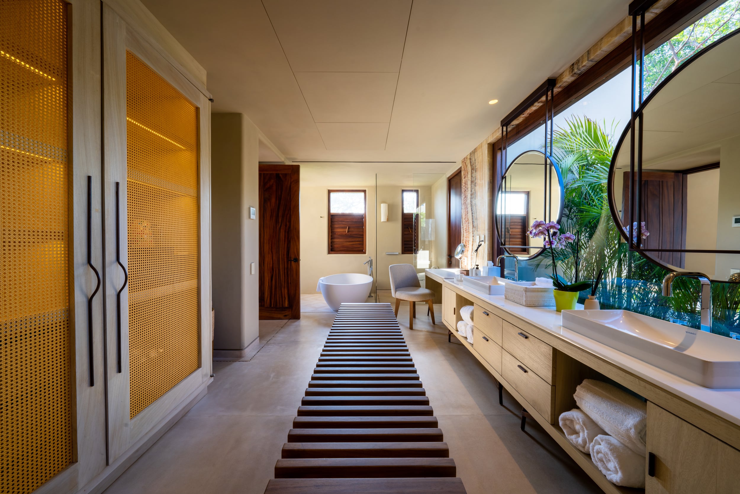 Wide primary ensuite bathroom with a freestanding oval soaking tub at the far end, a double-vanity with two vessel sinks and a striped runner on the floor, backlit amber cane wardrobe panels along the left wall, and a large arched mirror reflecting garden foliage through a window, Casa Akama, Ranchos Estates, Punta Mita.