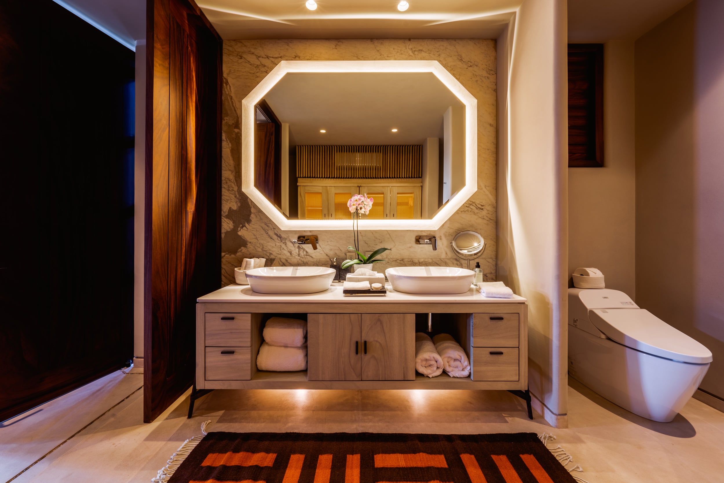 Ensuite bathroom with dual vessel sinks on a wood-drawer vanity, two round back-lit octagonal mirrors above a textured stone accent wall, folded towels on lower shelves, a Toto smart toilet to the right, and an orange-and-brown striped kilim rug on the tile floor, Casa Akama, Ranchos Estates, Punta Mita.