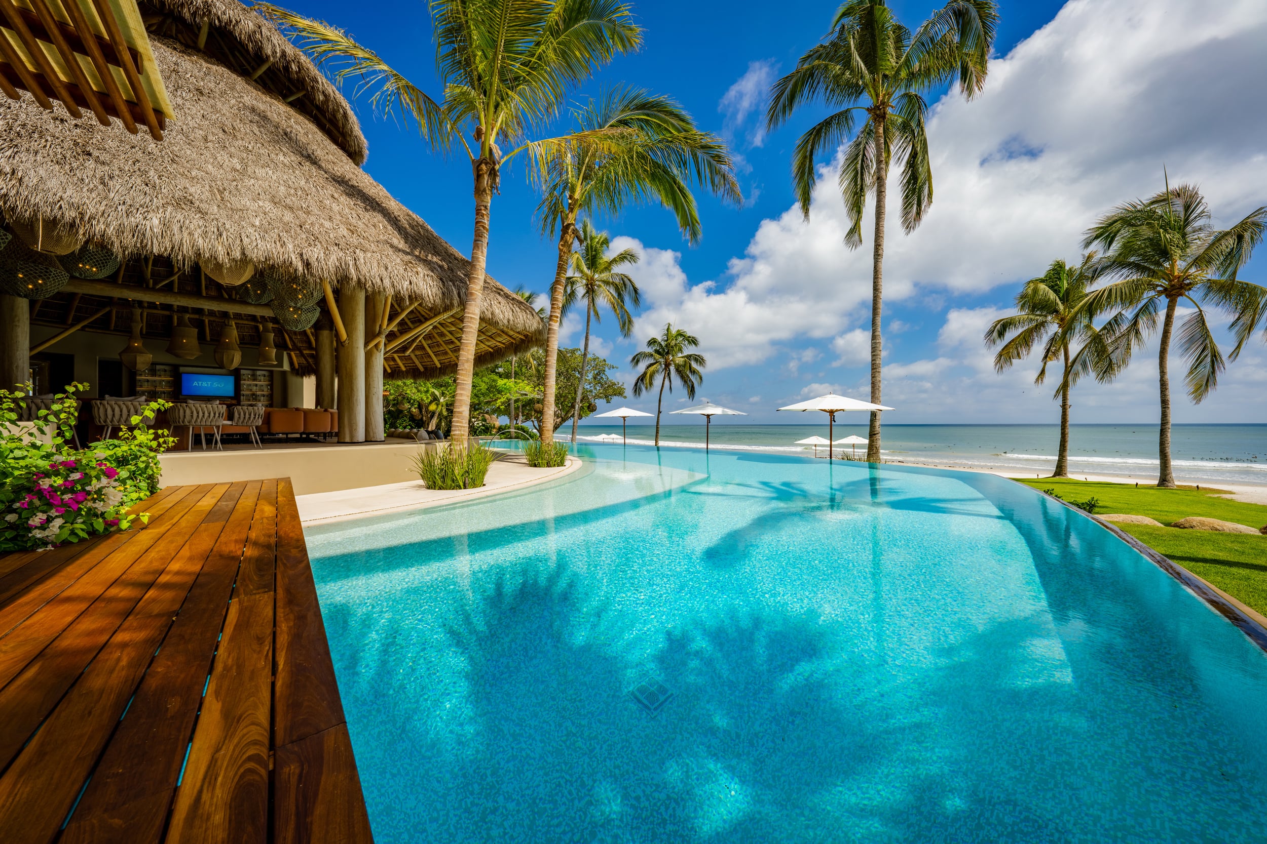 Daytime view of the main infinity pool from the hardwood deck level, with the thatched palapa pavilion on the left housing a bar and lounge, white market umbrellas spaced along the beach, coconut palms throughout, and the Pacific Ocean in the background under a blue sky, Casa Akama, Ranchos Estates, Punta Mita.