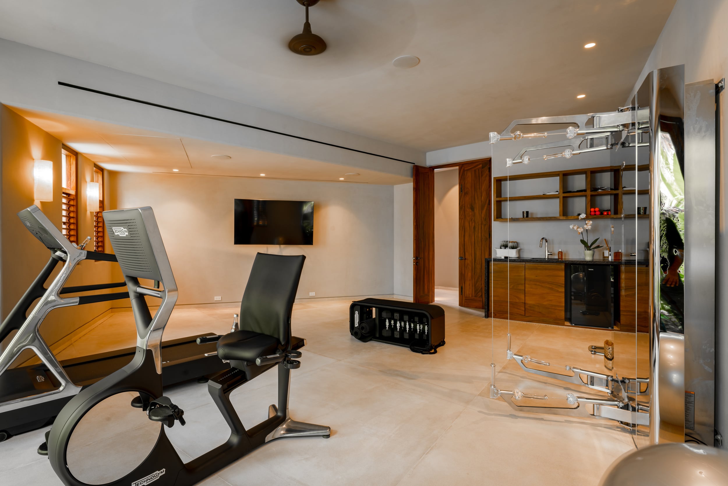 Indoor fitness room showing a treadmill and an adjustable seat exercise bike in the foreground, a wall-mounted television, a mini-bar cabinet with glassware on open shelves in the background, and a glass panel partition reflecting equipment, Casa Akama, Ranchos Estates, Punta Mita.