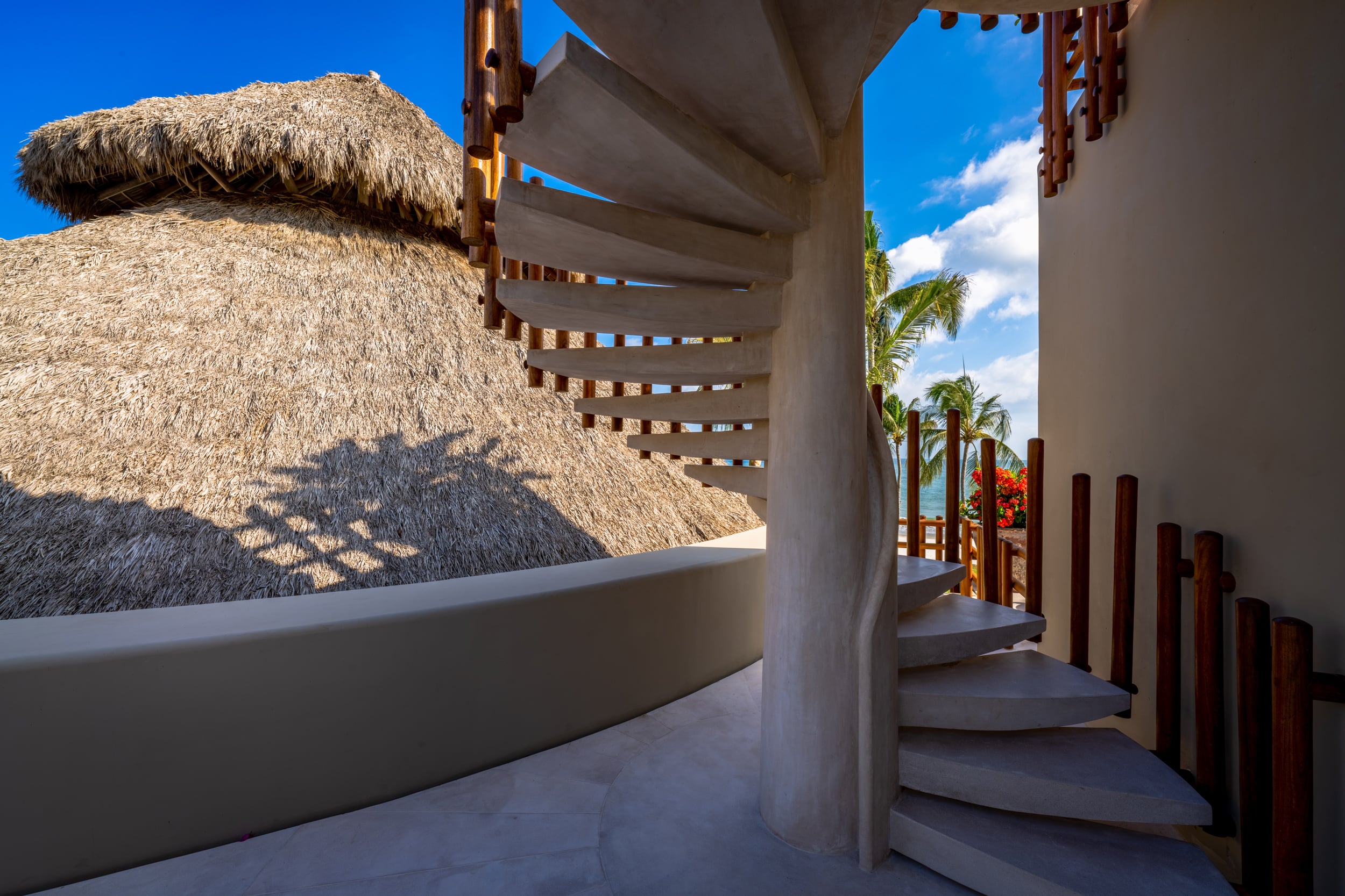 Outdoor concrete spiral staircase wrapping around a cylindrical white stucco column, adjacent to the edge of a thatched palapa roof with dappled shadow patterns, opening at the top to a view of palm trees, bougainvillea, and the Pacific Ocean under a clear blue sky, Casa Akama, Ranchos Estates, Punta Mita.