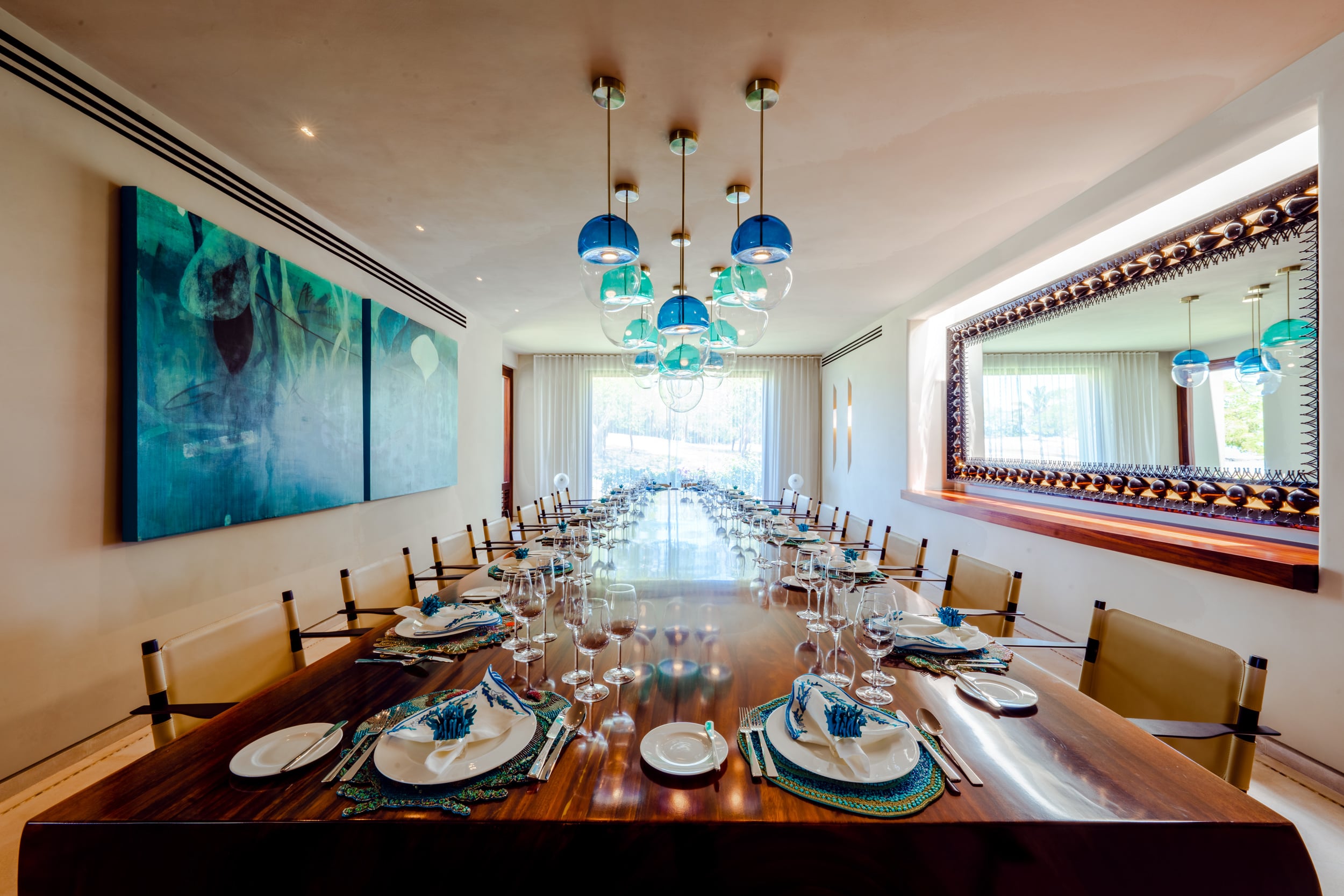 Formal dining room set for twenty with a long dark-wood table, rattan-back chairs, blue and teal blown-glass pendant lights overhead, a large triptych blue artwork on the left wall, and an ornate carved mirror in an alcove on the right, Casa Akama, Ranchos Estates, Punta Mita.