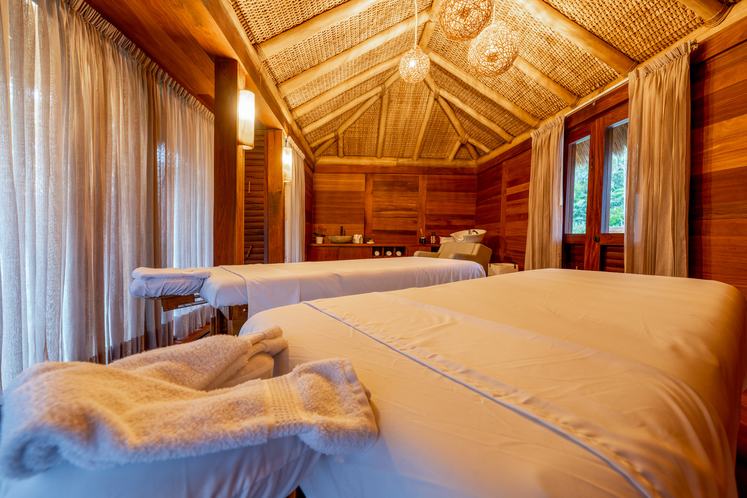 Dual-treatment massage room with vaulted ceiling clad in woven rattan panels, two massage tables made up in white linen with towels folded at the foot of each, cedar-plank walls, woven pendant lights, and sheer curtains filtering garden light from both sides, Casa Akama, Ranchos Estates, Punta Mita.