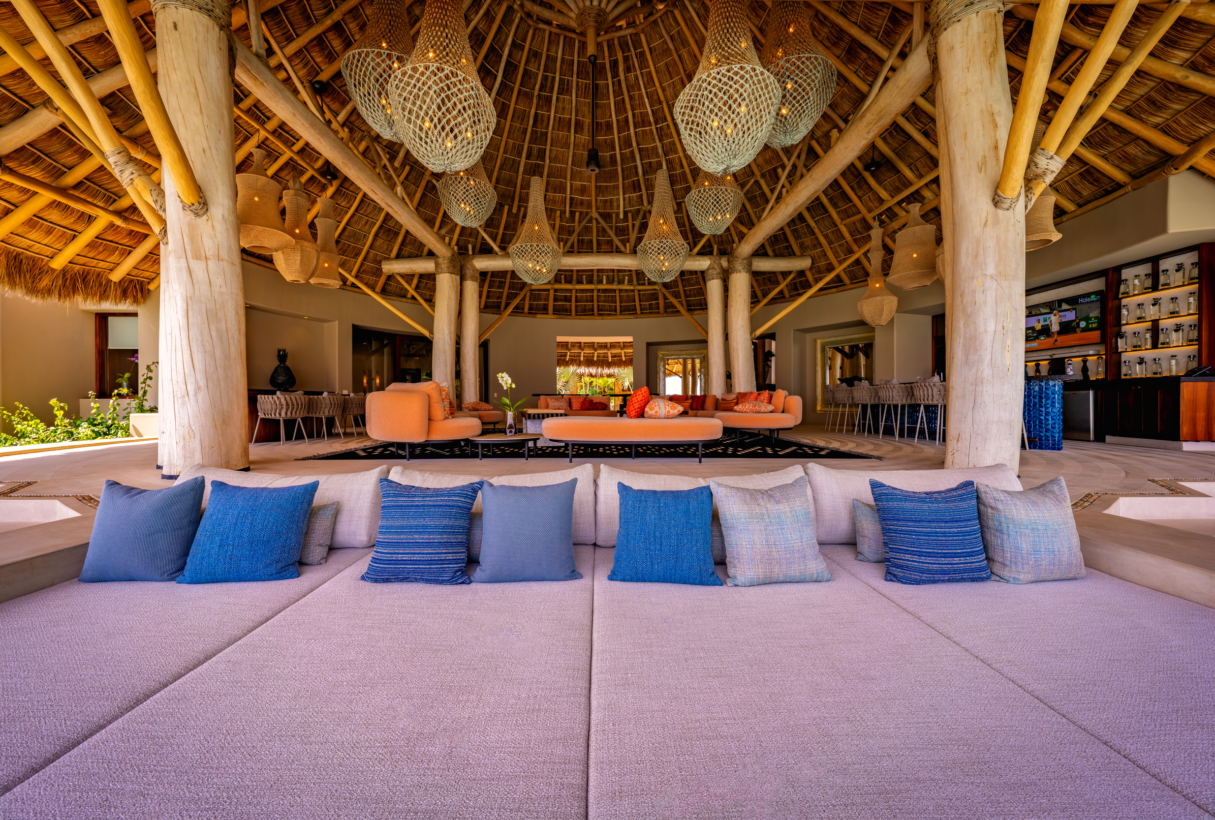 Open-air palapa lounge interior viewed from a wide day-bed with blue and grey cushions in the foreground, looking toward a cluster of woven pendant chandeliers, polished timber columns, orange curved sofas, and a bar at right, all beneath a high thatched roof, Casa Akama, Ranchos Estates, Punta Mita.