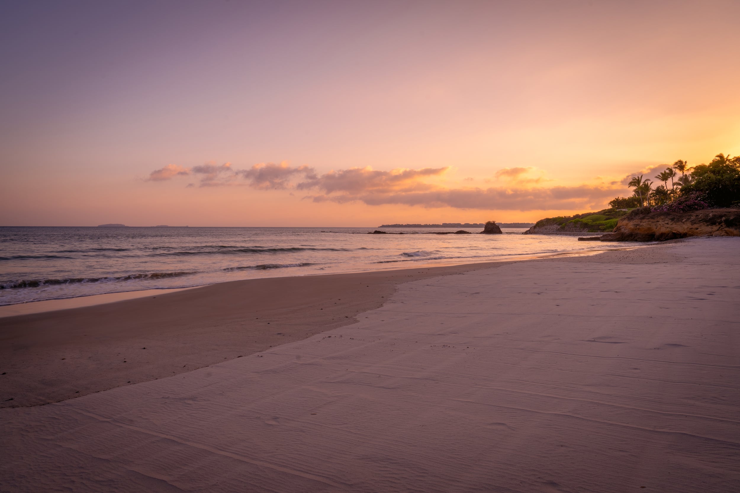 Private beach at dusk with wide flat pale sand, gentle surf, a rocky outcrop at the waterline, and palms with bougainvillea on the headland to the right under a gradient sky of orange and lavender, Casa Akama, Ranchos Estates, Punta Mita.
