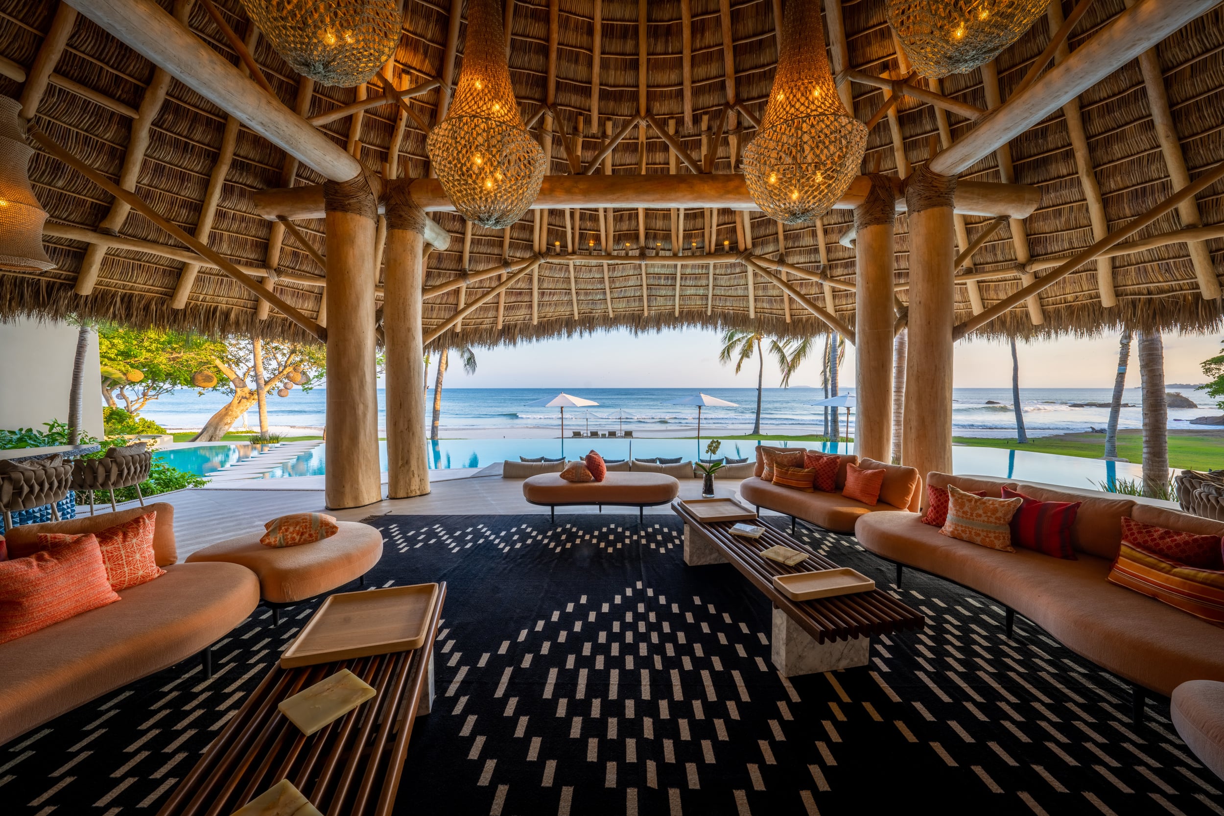 Open-air palapa living pavilion at dusk with curved terracotta-tone sofas, a geometric black-and-white rug, low coffee tables, woven pendant lights overhead, and an unobstructed view through the timber columns to the pool, beach, and Pacific Ocean beyond, Casa Akama, Ranchos Estates, Punta Mita.