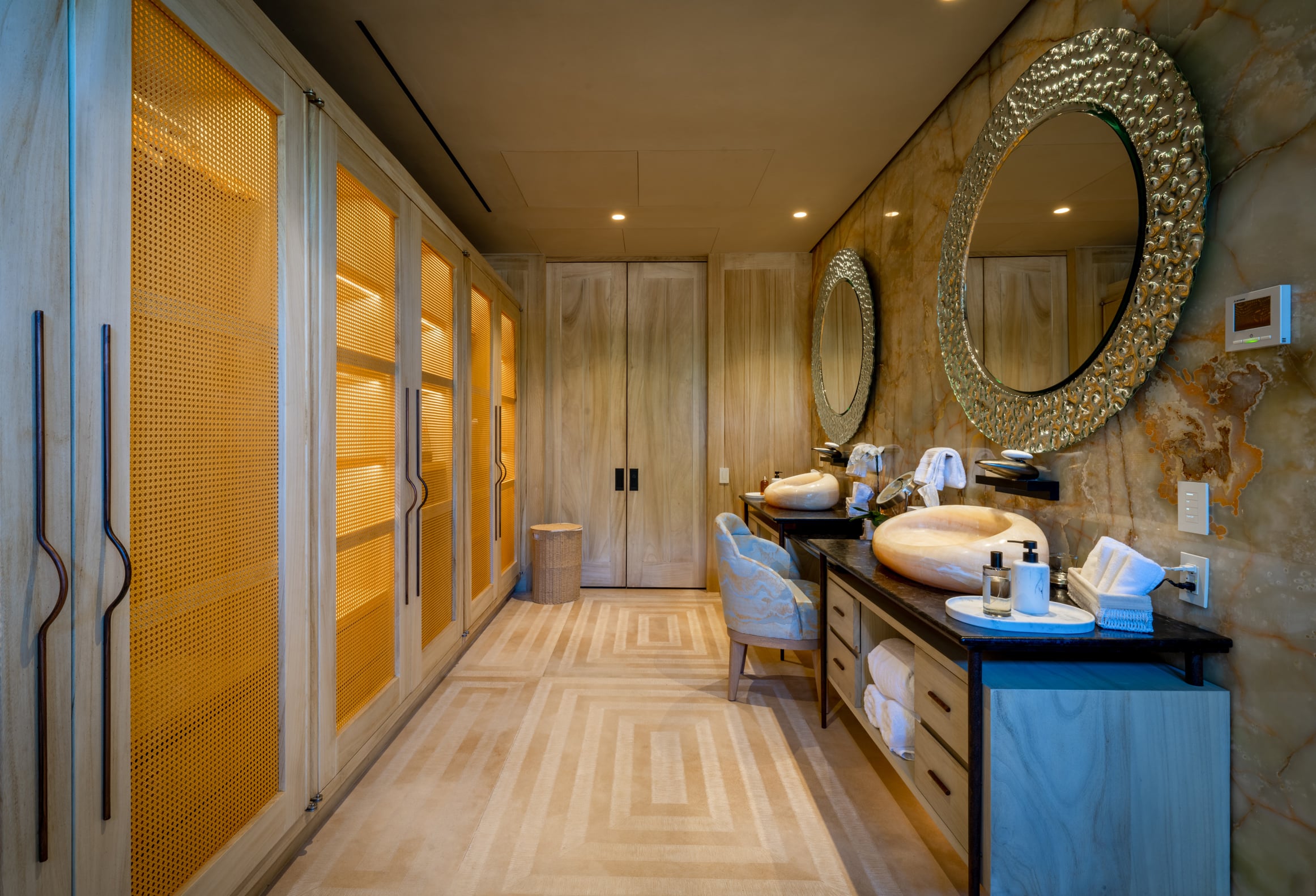 Ensuite dressing corridor with backlit amber-mesh wardrobe cabinets along the left wall, a herringbone-tile floor, and a double vanity with two stone vessel sinks on a dark counter topped by two ornate silver-leafed circular mirrors with textured frames, set against an onyx-veined wall, Casa Akama, Ranchos Estates, Punta Mita.