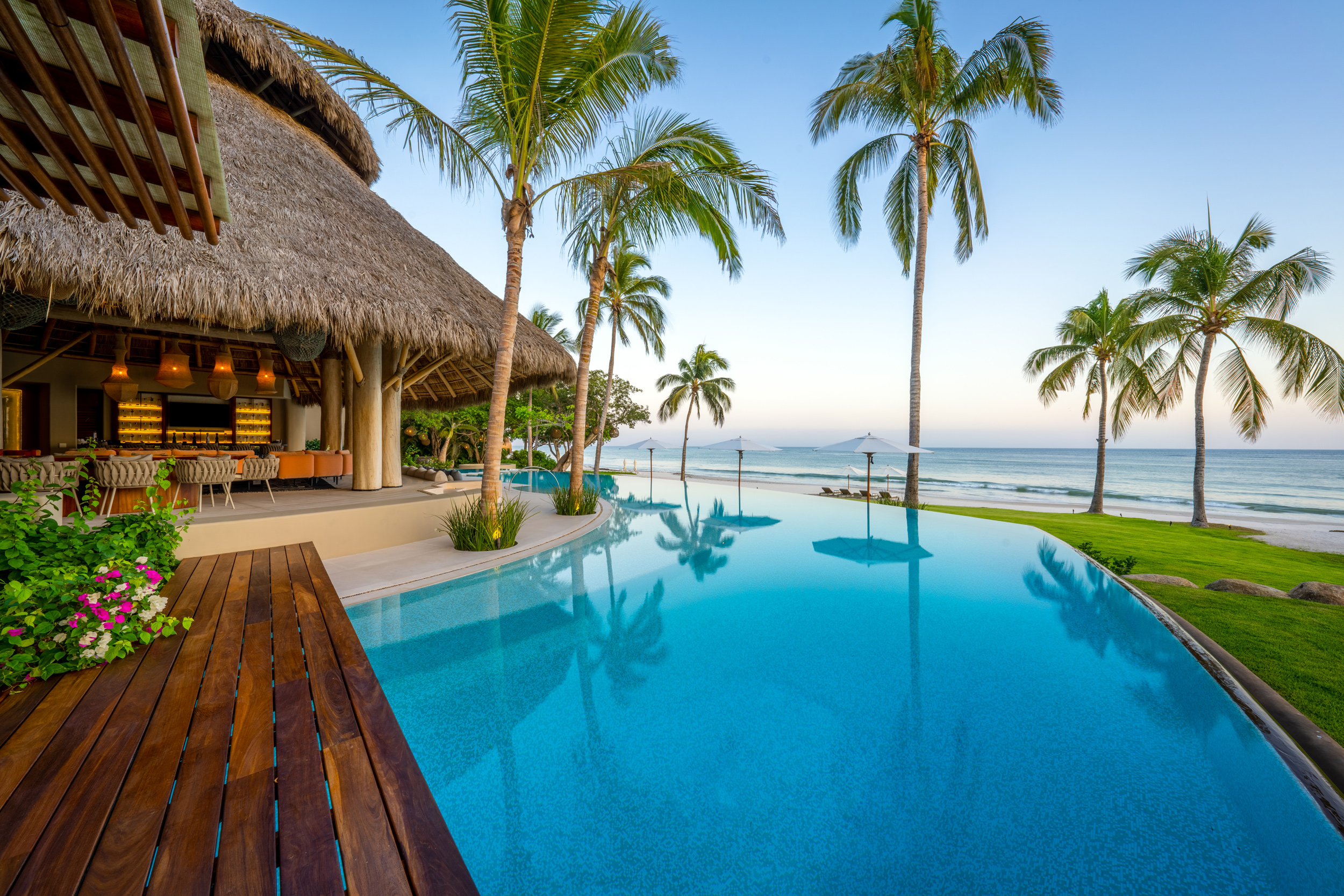 Infinity pool at early evening reflecting a pastel sky, bordered by a hardwood deck with bougainvillea in the foreground, the palapa bar pavilion to the left under warm internal lighting, coconut palms framing the pool lane, and the Pacific Ocean visible to the right at twilight, Casa Akama, Ranchos Estates, Punta Mita.