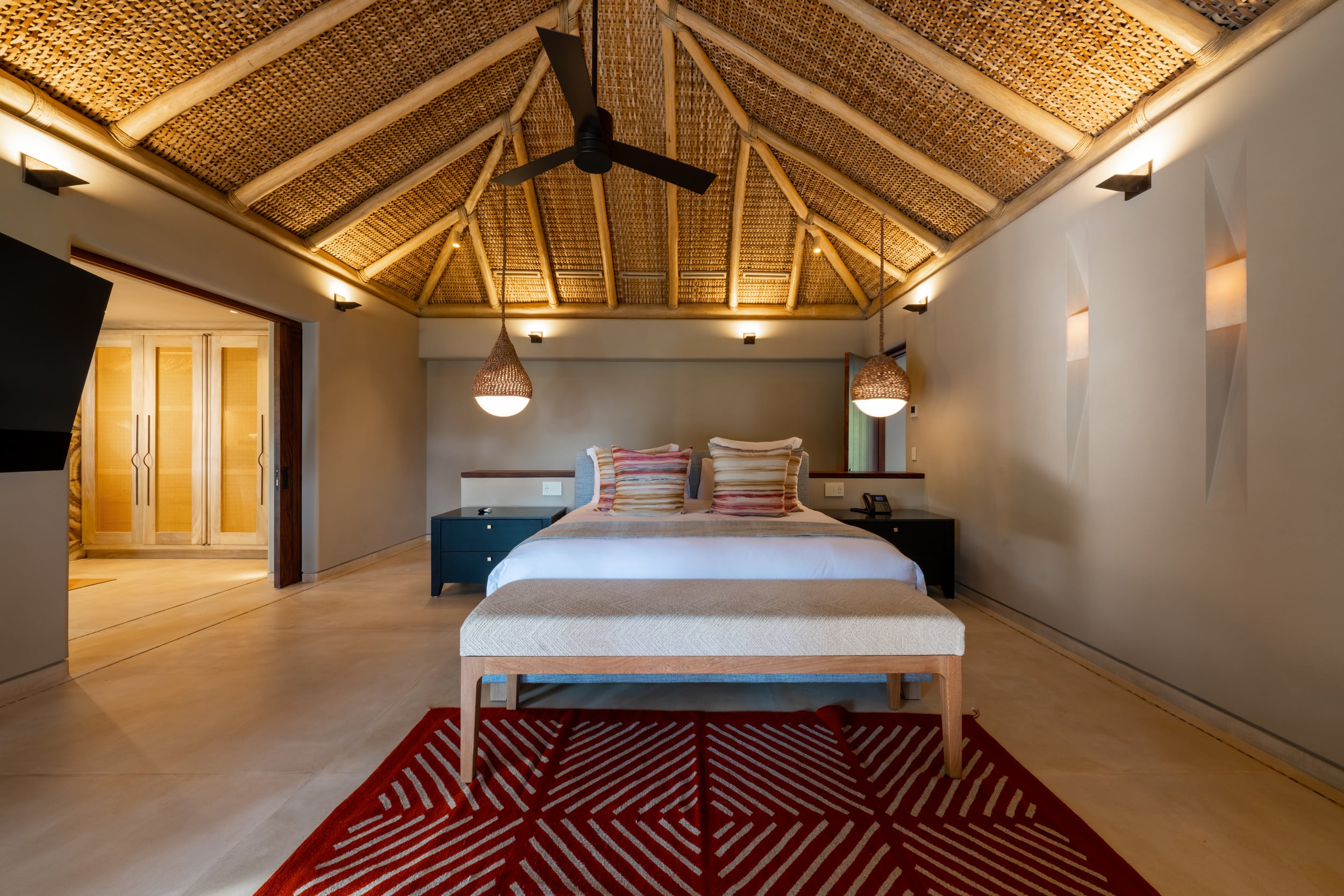 Guest bedroom with vaulted rattan ceiling lit by warm uplighting, a king bed with a striped red-ground kilim rug at its foot, two hanging wicker pendant lamps, a wooden bench, and backlit amber cane wardrobe doors visible through a doorway at left, Casa Akama, Ranchos Estates, Punta Mita.