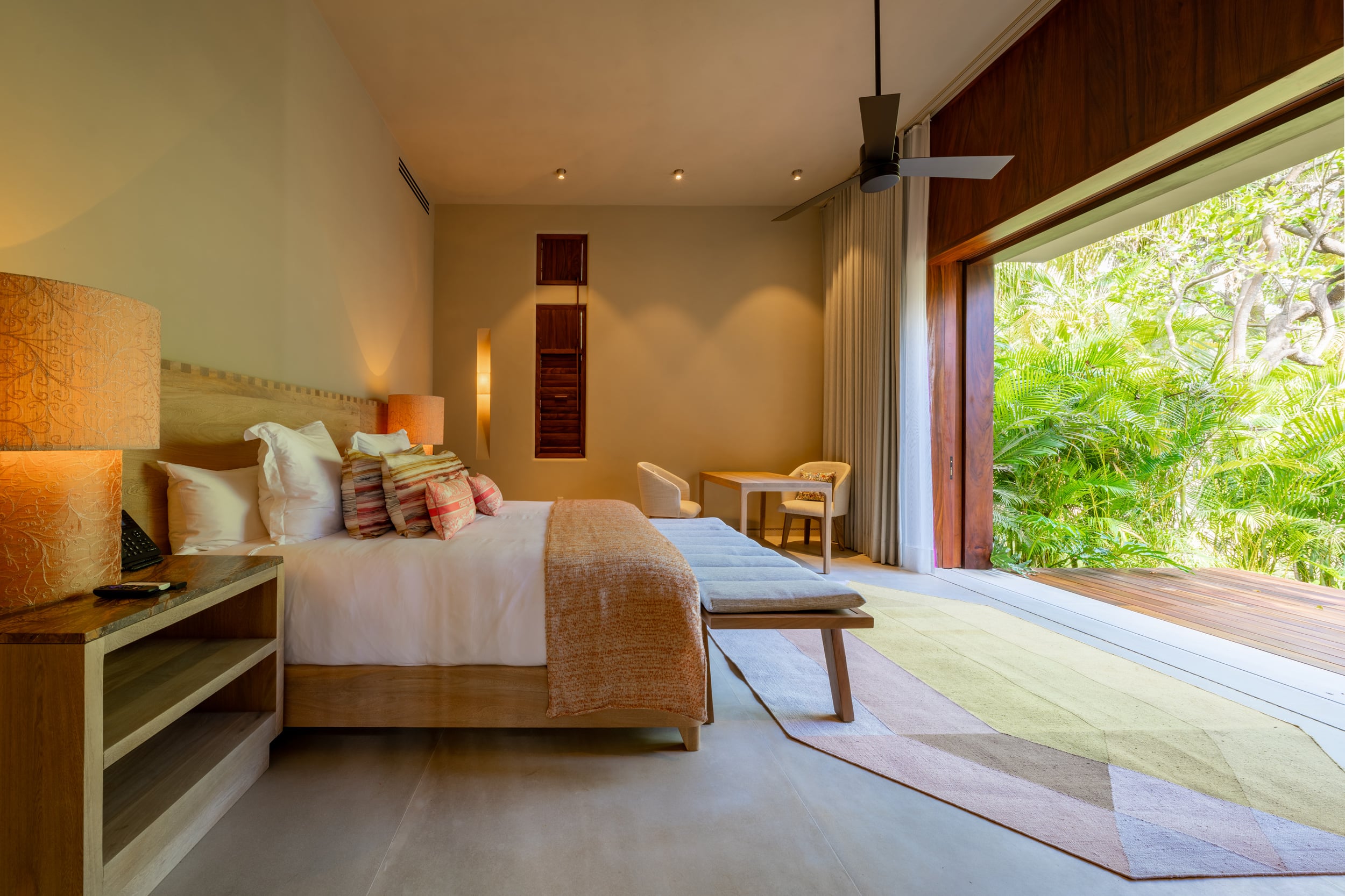 Guest bedroom with an orange bedside lamp, a king bed dressed in white linen with multicolor ikat accent pillows, a padded bench at the foot, and a wide sliding door standing fully open to a terrace with dense garden planting and dappled afternoon light, Casa Akama, Ranchos Estates, Punta Mita.