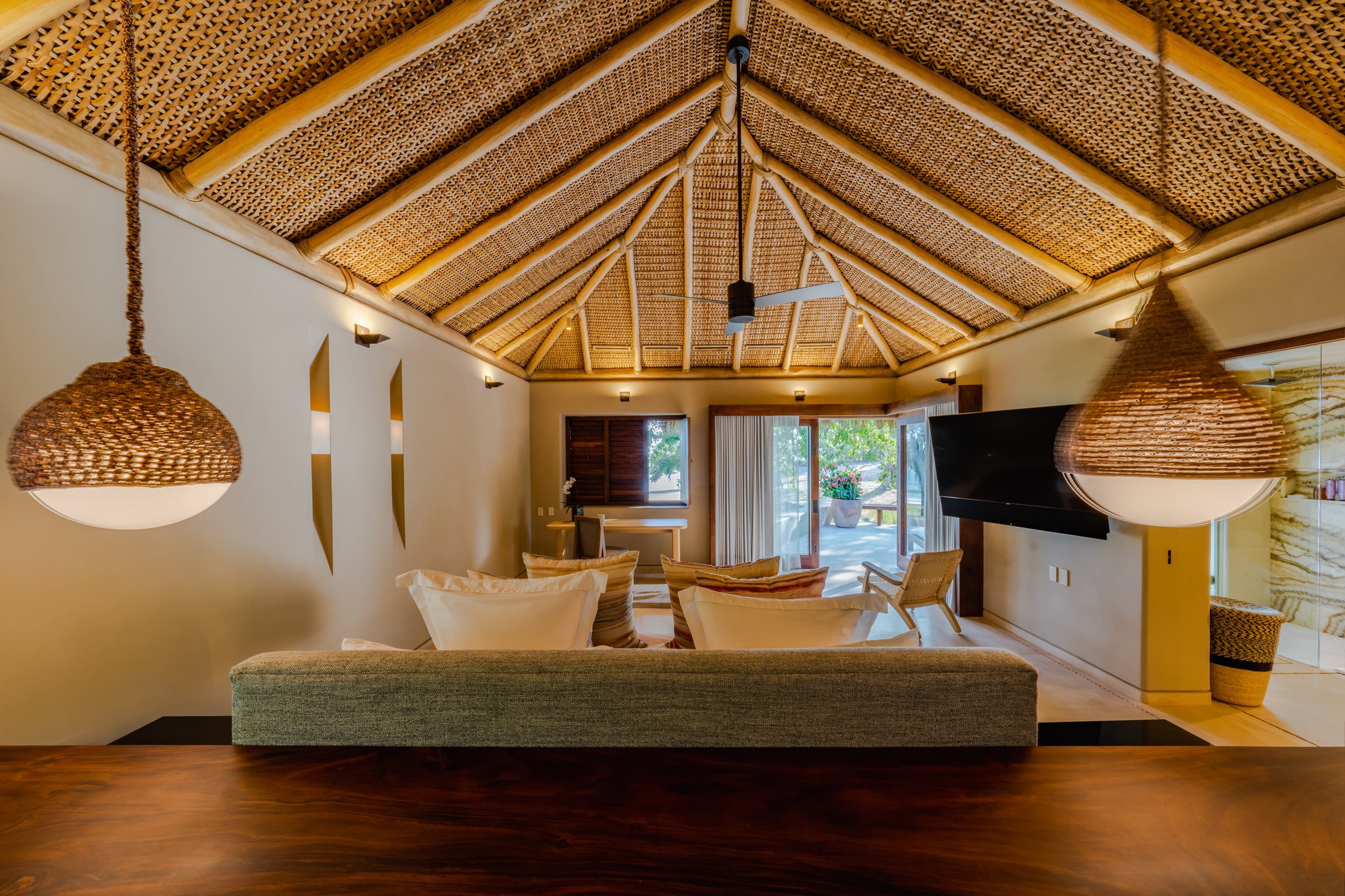 Interior sitting room with a vaulted woven-cane ceiling, a long low grey sofa, woven globe pendant lamps on each side, a wall-mounted flat-screen television, and glass doors at the rear opening to a garden terrace, the room finished with a dark polished concrete floor, Casa Akama, Ranchos Estates, Punta Mita.