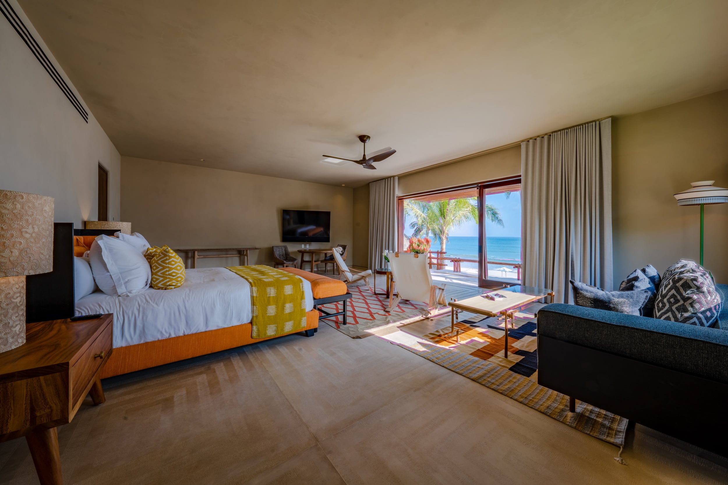 Spacious beachfront guest room with a king bed dressed in orange and yellow ikat textiles, a sitting area of canvas sling chairs and a dark sofa, a wall-mounted television, and large sliding glass doors fully open to a sun-flooded terrace overlooking the ocean and a coconut palm, Casa Akama, Ranchos Estates, Punta Mita.