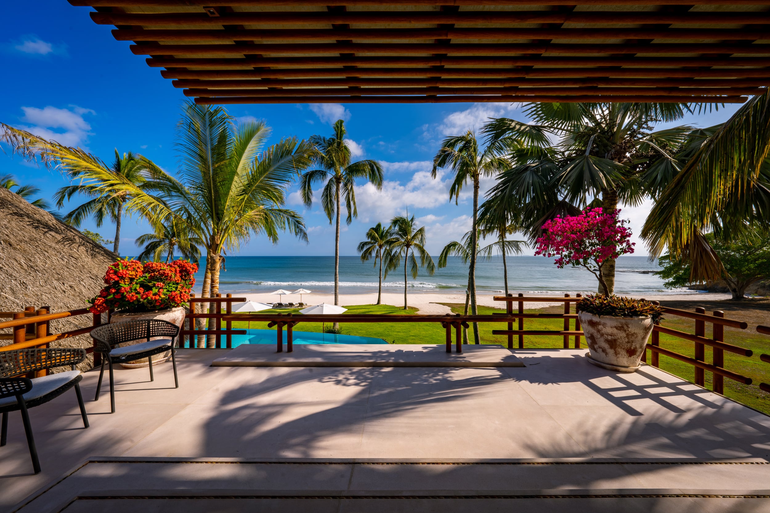 Covered terrace with a slatted wood pergola overhead, looking out past flowering bougainvillea and red geraniums toward the pool, a row of coconut palms, the beach, and open Pacific horizon under a bright blue sky with a large ceramic planter on the right, Casa Akama, Ranchos Estates, Punta Mita.