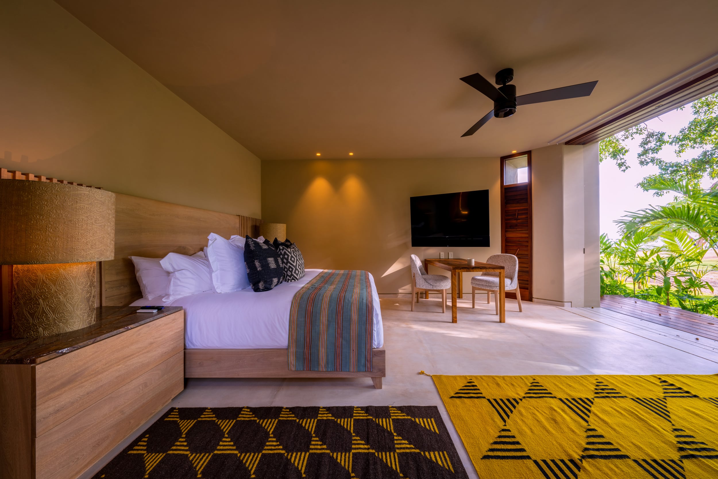 Guest bedroom with a king bed on a natural-wood platform, dark indigo and yellow geometric rug, warm recessed ceiling lighting, a flat-screen television on the wall, a small work table and chairs, and wide sliding doors fully open to a garden terrace, Casa Akama, Ranchos Estates, Punta Mita.