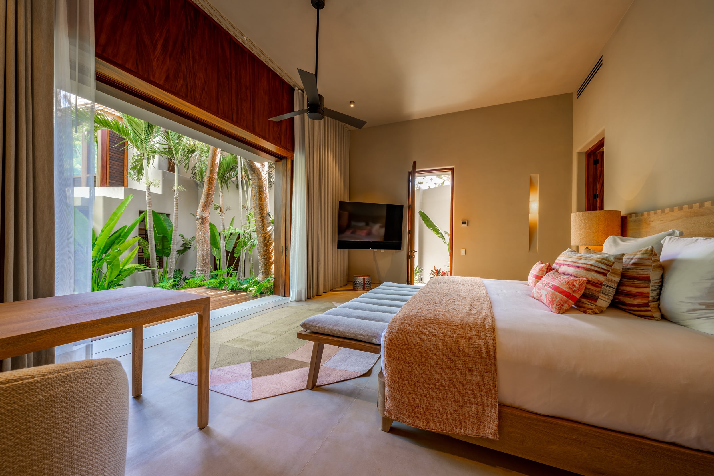 Guest bedroom with a king bed dressed in blush linen and ikat cushions, a padded bench at the foot, a wall-mounted television, and a full-width pivoting glass door open to a private plunge-pool courtyard with dense tropical planting visible outside, Casa Akama, Ranchos Estates, Punta Mita.