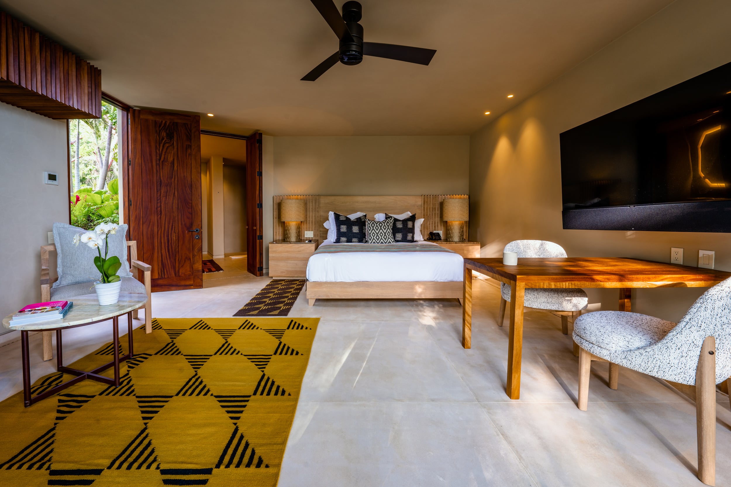 Guest bedroom with a king bed on a light-oak headboard, black and ivory ikat accent pillows, a yellow and black geometric rug, a wood worktable and upholstered chairs, wall-mounted television, and garden light entering through a louvered side door, Casa Akama, Ranchos Estates, Punta Mita.