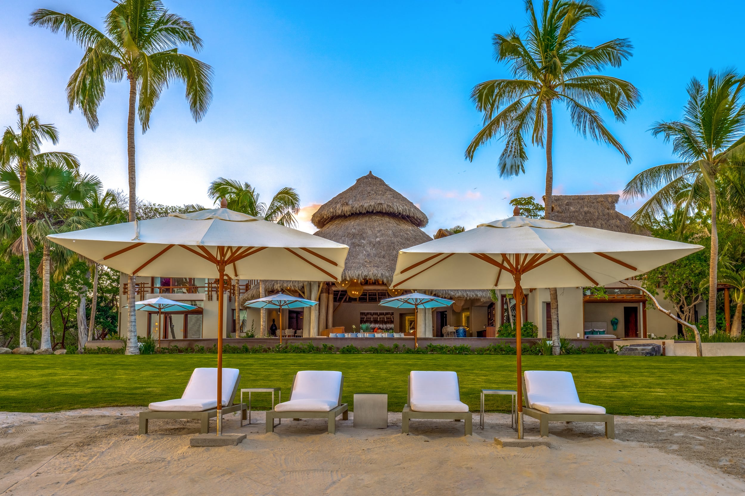 View from the beach looking back at the villa compound at dusk, showing four white market umbrellas with sun loungers on pale sand, a manicured lawn rising to the main palapa-roofed pavilion flanked by coconut palms, under a deep blue evening sky, Casa Akama, Ranchos Estates, Punta Mita.