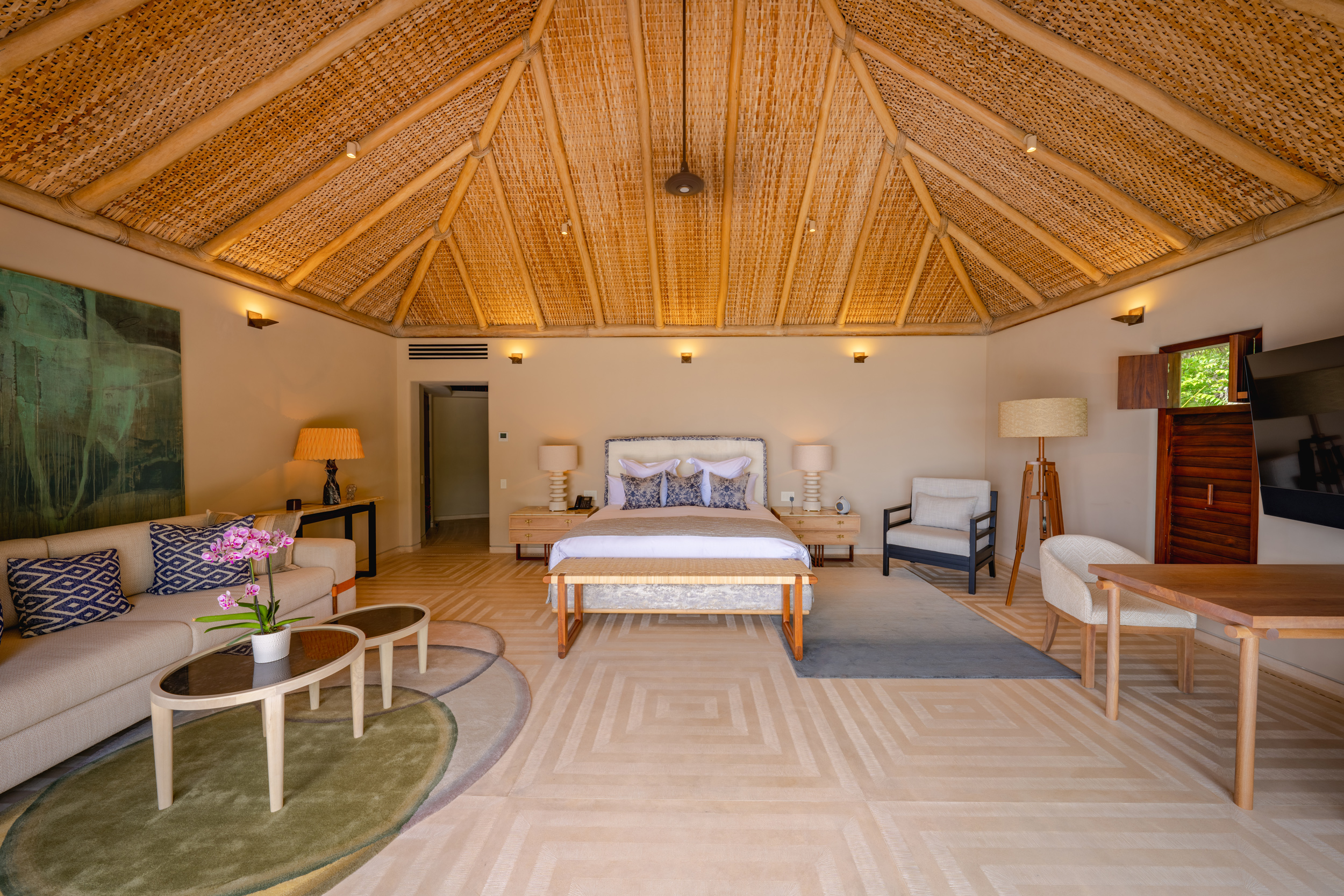 Vaulted palapa suite with woven reed ceiling, king bed centered on herringbone-tile floor, sitting area with sofa and orchid-topped coffee table, and large canvas art on the left wall, Casa Akama, Ranchos Estates, Punta Mita.