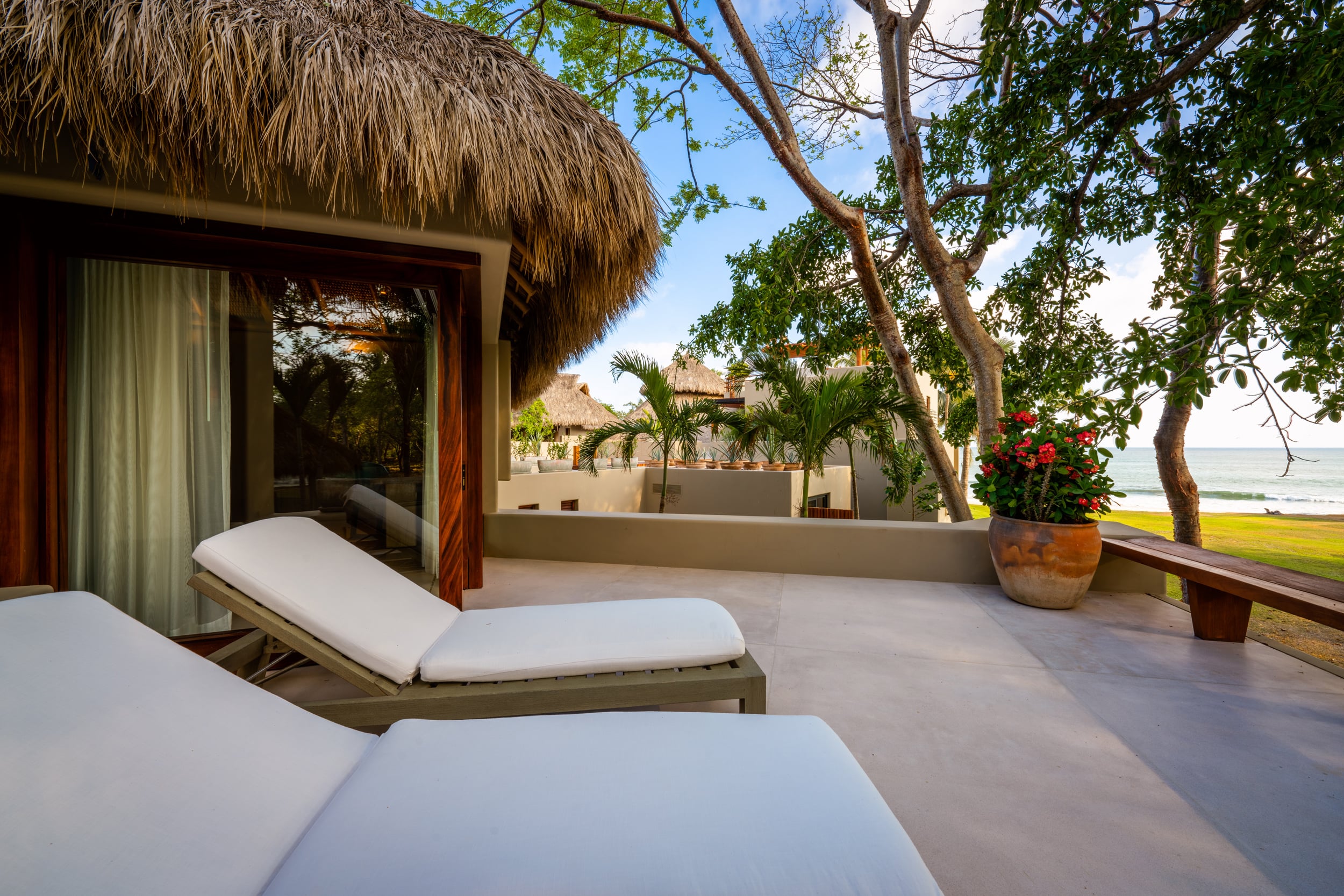 Private terrace off a palapa-roofed suite with two cushioned lounge chairs, terracotta pot of red bougainvillea, and unobstructed view of the Pacific Ocean through tropical trees, Casa Akama, Ranchos Estates, Punta Mita.