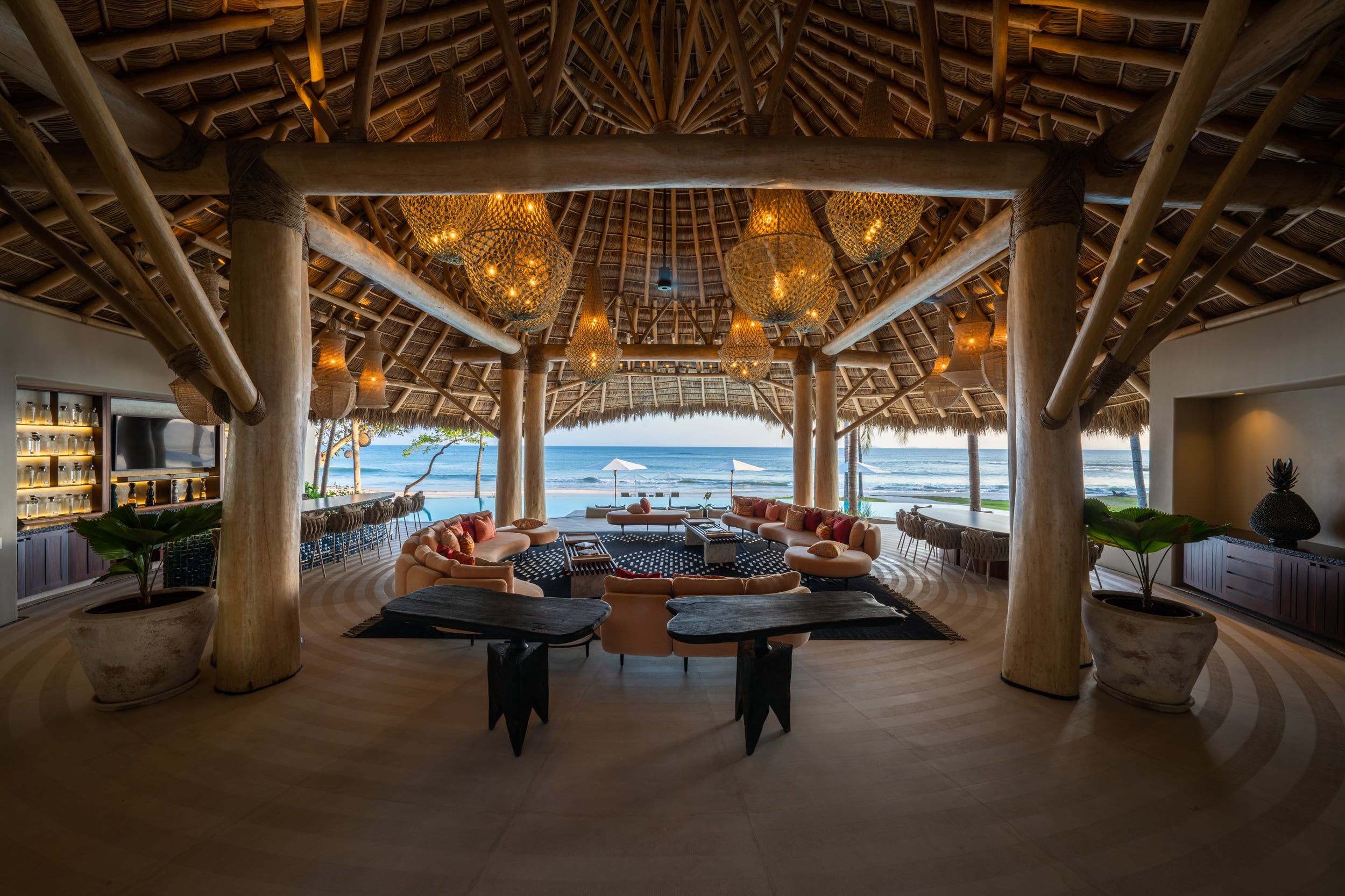 Open-air living pavilion with tall palapa roof supported by thick timber columns, cluster of woven-globe pendant lights, curved sectional sofas in terracotta tones, bar shelving on the left, and a full ocean horizon framed by the open facade, Casa Akama, Ranchos Estates, Punta Mita.