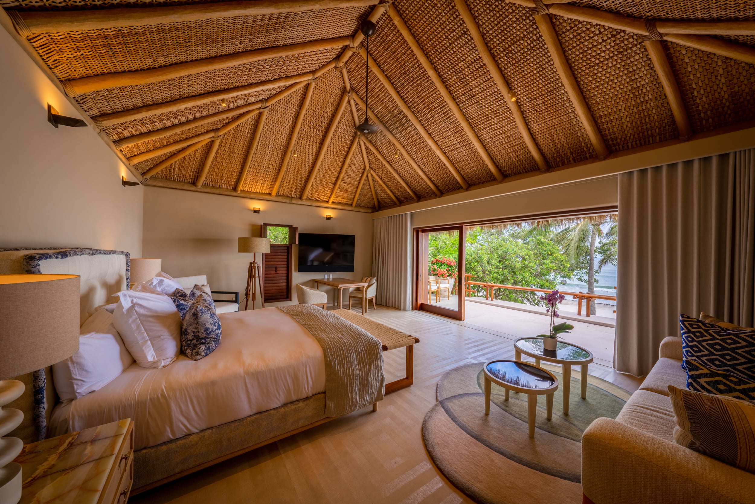 King bedroom under a high woven palapa roof with sliding glass doors open to a balcony overlooking palm trees and the ocean, a sofa seating area, flat-screen television, and bedside lamps flanking the upholstered headboard, Casa Akama, Ranchos Estates, Punta Mita.