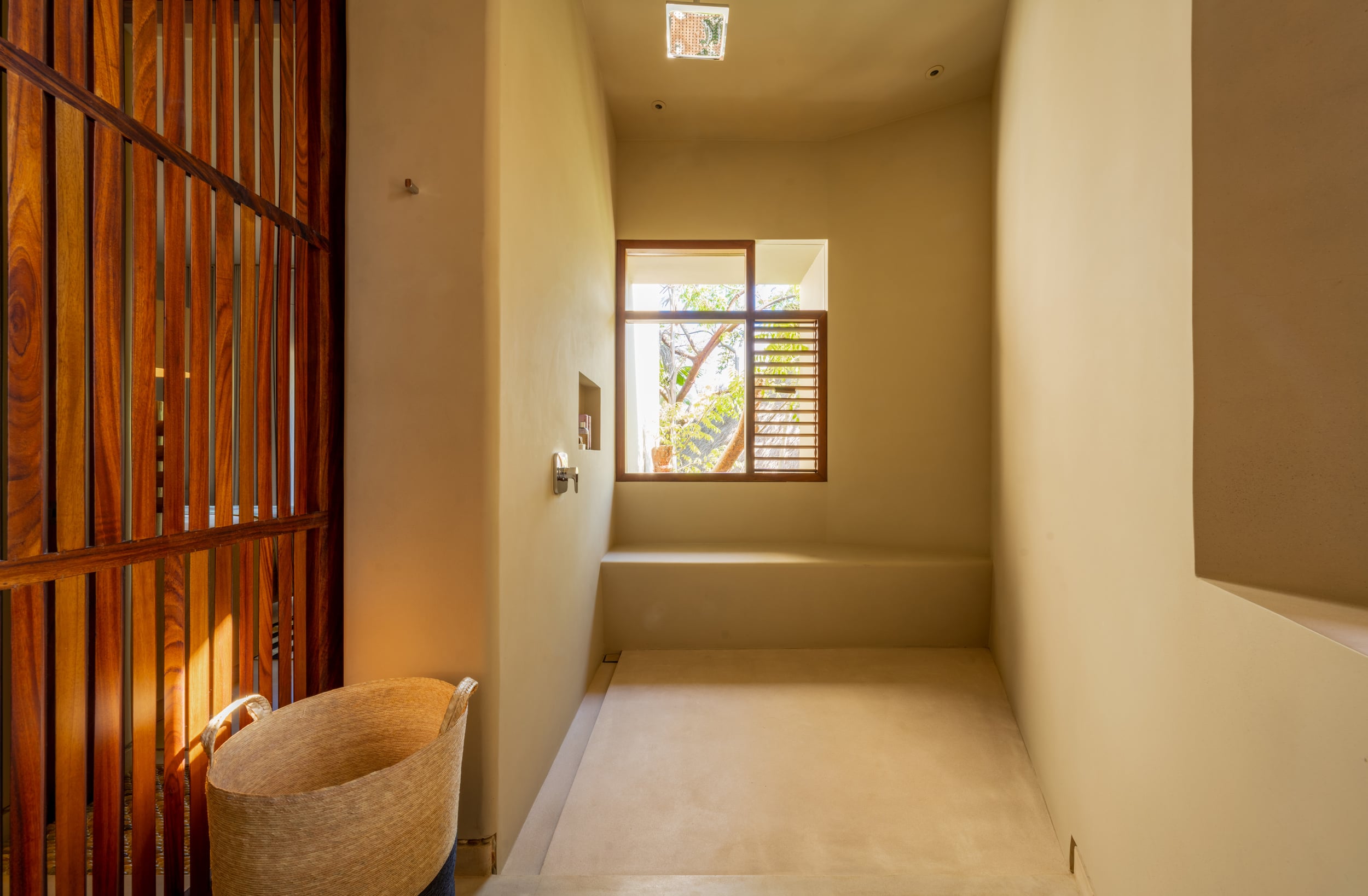 Sunlit bathroom passage with warm plaster walls, wood-slat partition gate on the left, a recessed niche, louvered window admitting natural light, and a step-down soaking area at the far end with a woven basket on the floor, Casa Akama, Ranchos Estates, Punta Mita.