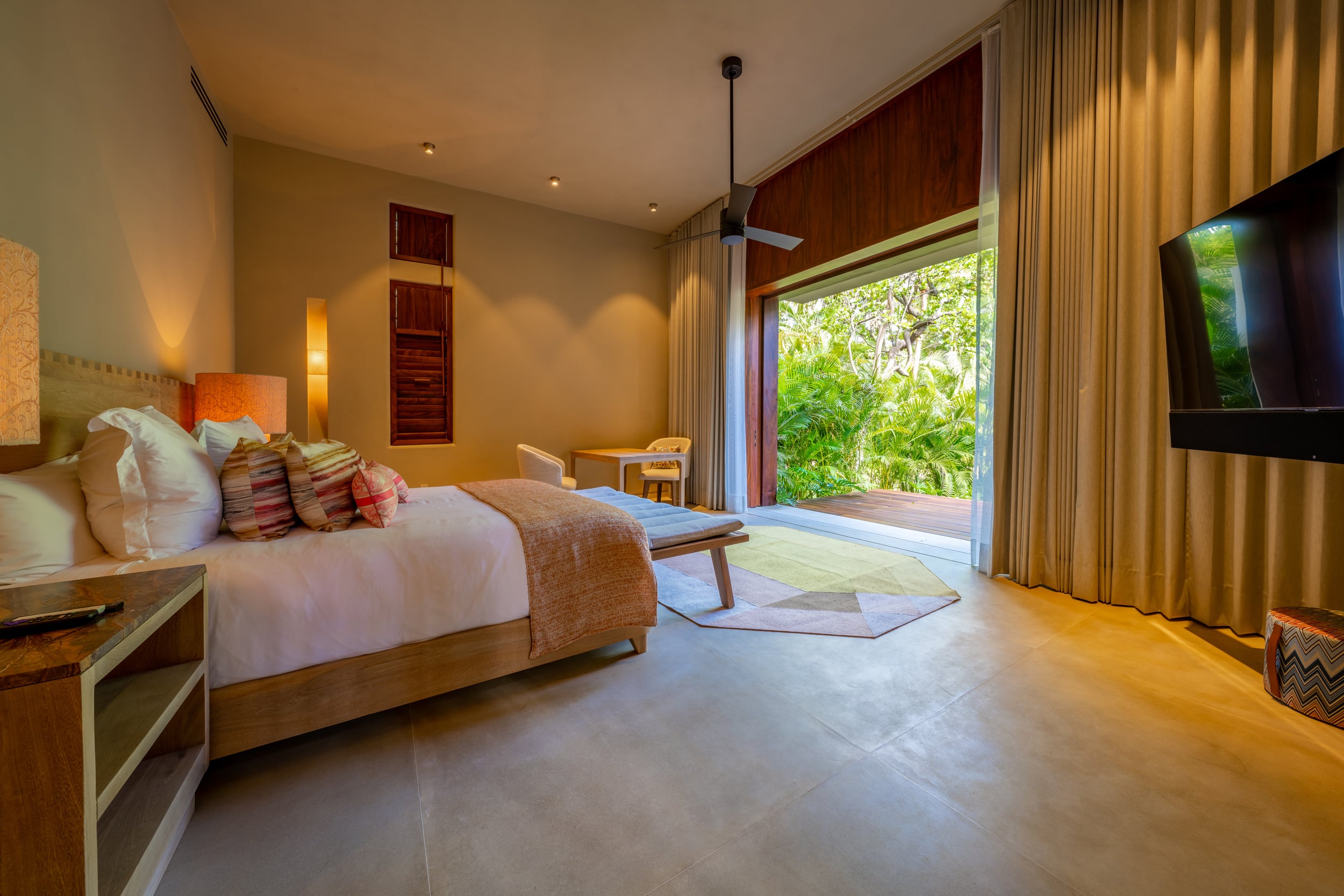 Guest bedroom with concrete floor, king bed dressed in warm-toned linens and striped throw pillows, ceiling fan, louvered wood shutters, flat-screen television, and sliding glass door open to a lush garden terrace, Casa Akama, Ranchos Estates, Punta Mita.