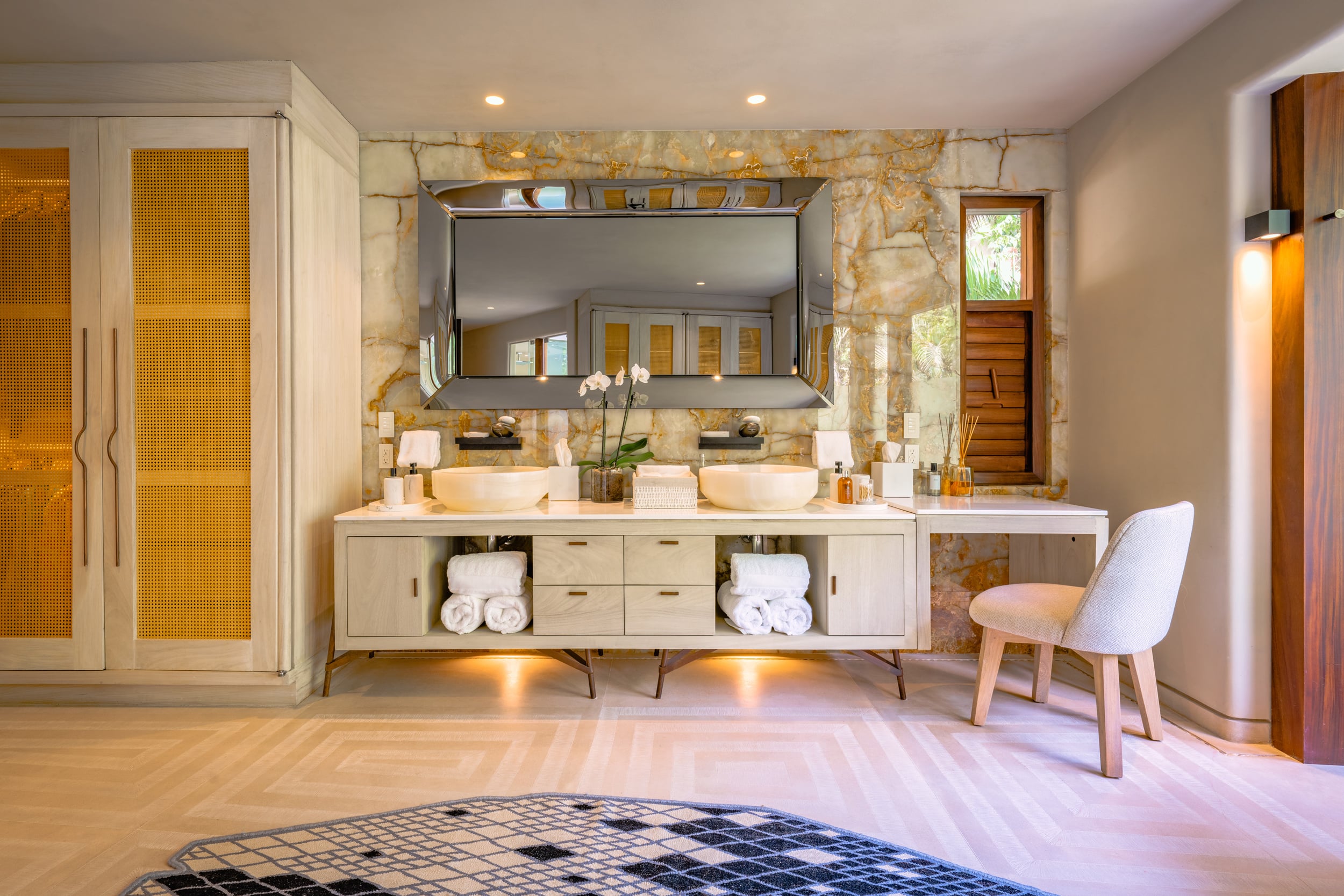 Ensuite bathroom vanity area with two vessel sinks on a cream floating console, broad stone-veneer mirror, amber onyx accent wall, rattan wardrobe doors on the left, and a vanity desk with white chair on the right, Casa Akama, Ranchos Estates, Punta Mita.