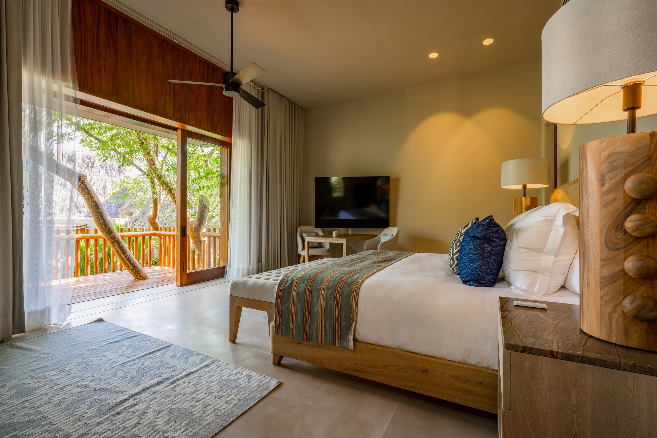 Guest bedroom with king bed, carved wood headboard, blue and teal accent pillow, sliding glass door open to a tree-shaded wooden deck, flat-screen television on a media unit, and a ceiling fan overhead, Casa Akama, Ranchos Estates, Punta Mita.