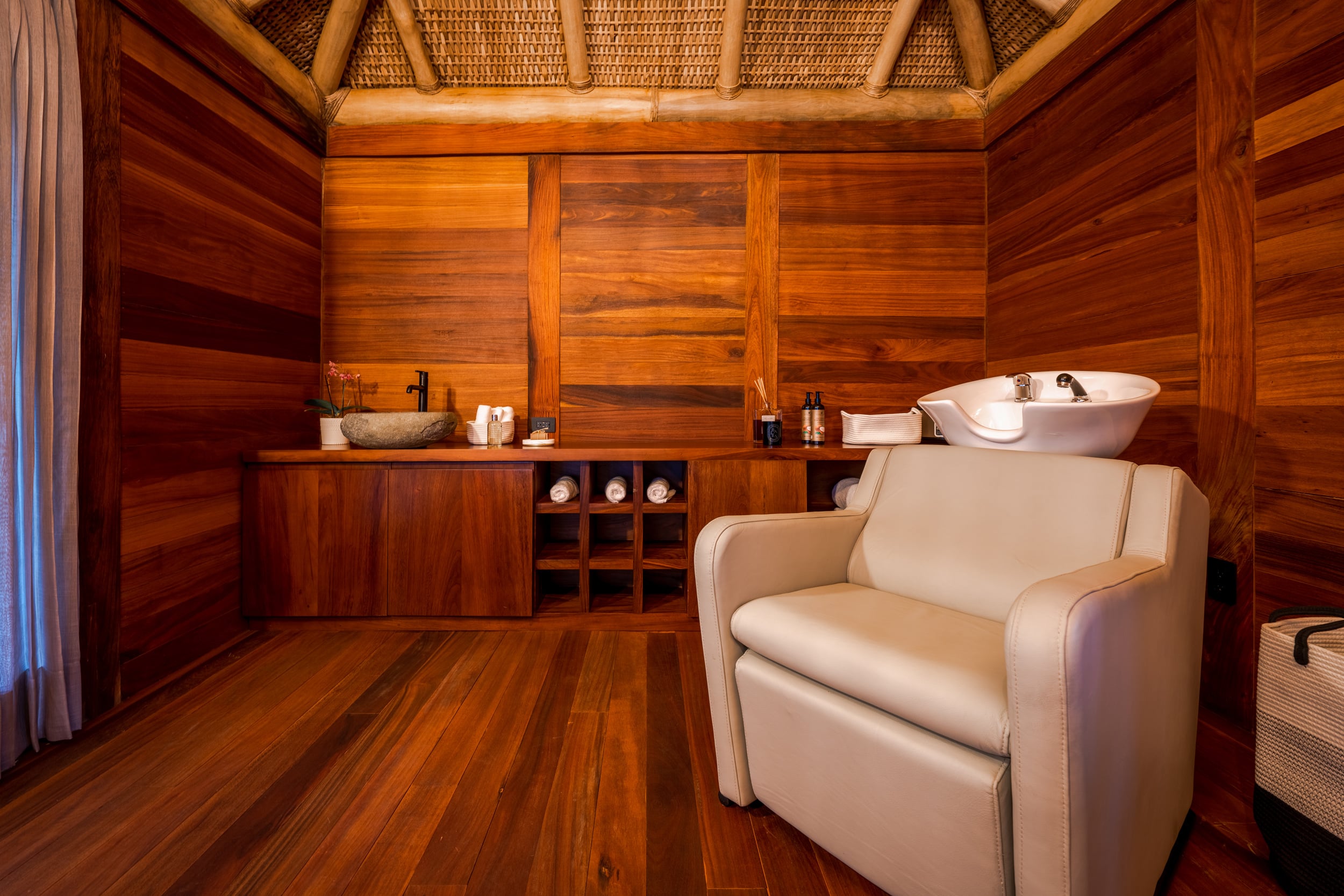 Spa treatment room lined with warm-toned hardwood planks on walls, ceiling, and floor, featuring a salon shampoo chair with white basin, a stone vessel sink on a low wood counter, and a small woven-reed palapa ceiling above, Casa Akama, Ranchos Estates, Punta Mita.