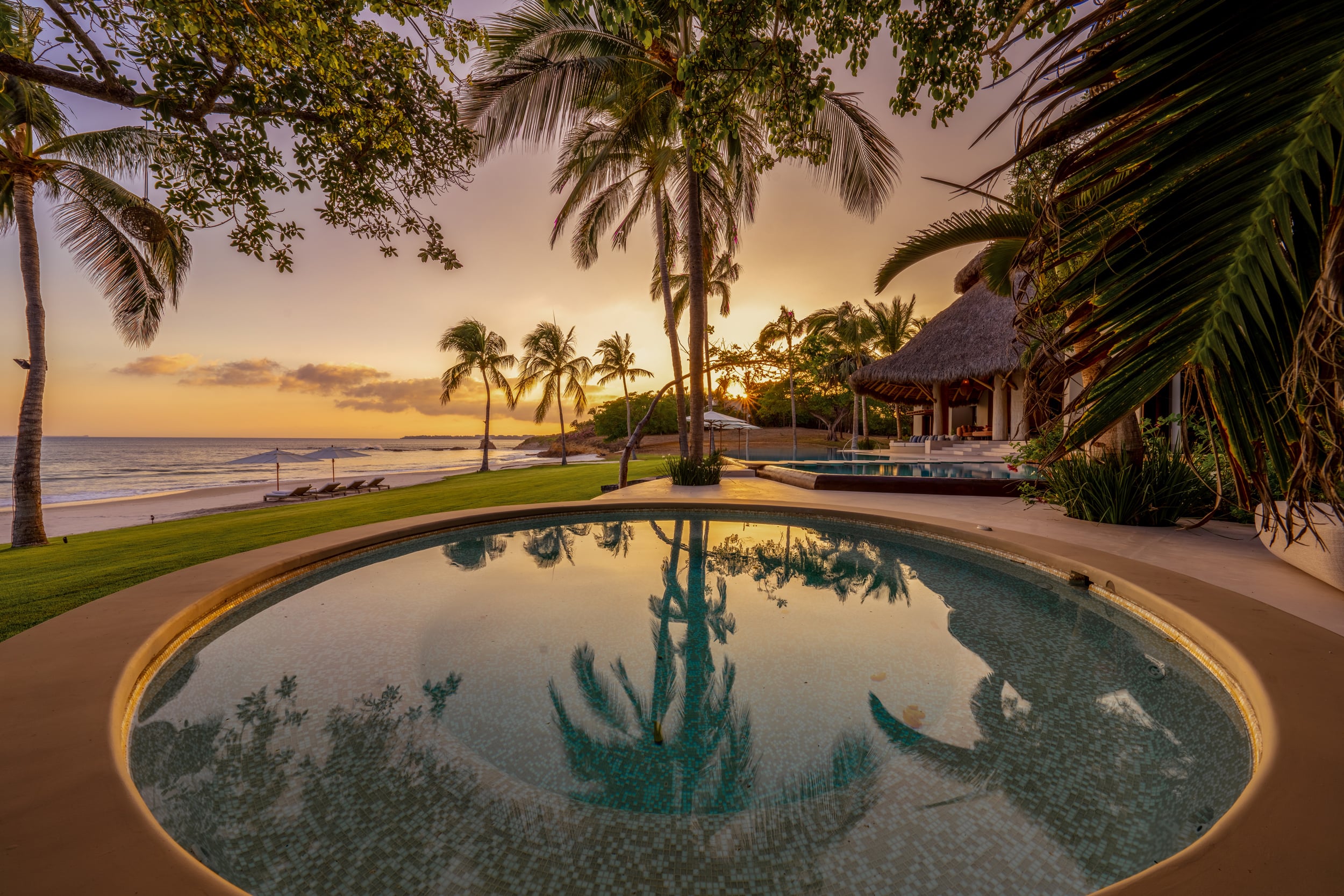 Circular plunge pool in the foreground reflecting palm fronds at sunset, with the main infinity pool, beach umbrellas, lawn, and the Pacific Ocean stretching to the horizon behind it, Casa Akama, Ranchos Estates, Punta Mita.
