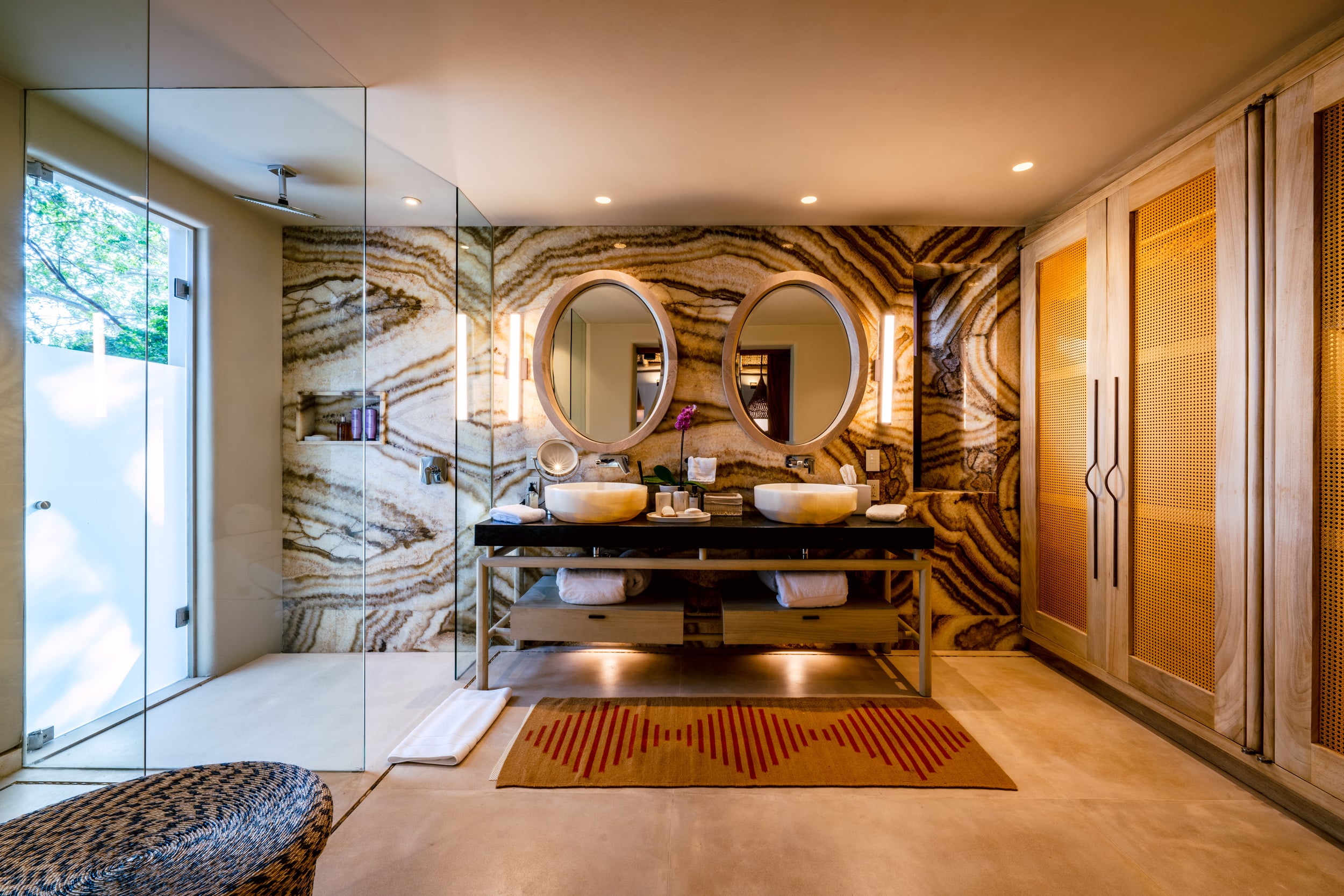 Primary ensuite bathroom with bold swirling agate-patterned stone wall, two round mirrors above a dark floating double vanity with vessel sinks, a glass-enclosed walk-in shower on the left, and rattan wardrobe doors on the right, Casa Akama, Ranchos Estates, Punta Mita.