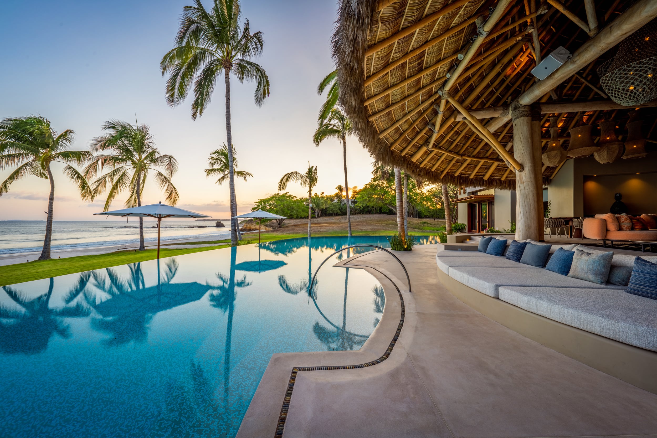 Infinity pool at dusk with a built-in daybed ledge under a palapa roof on the right, rows of palm trees reflected in still water, and beach umbrellas along the shoreline as the sky transitions from blue to warm gold, Casa Akama, Ranchos Estates, Punta Mita.
