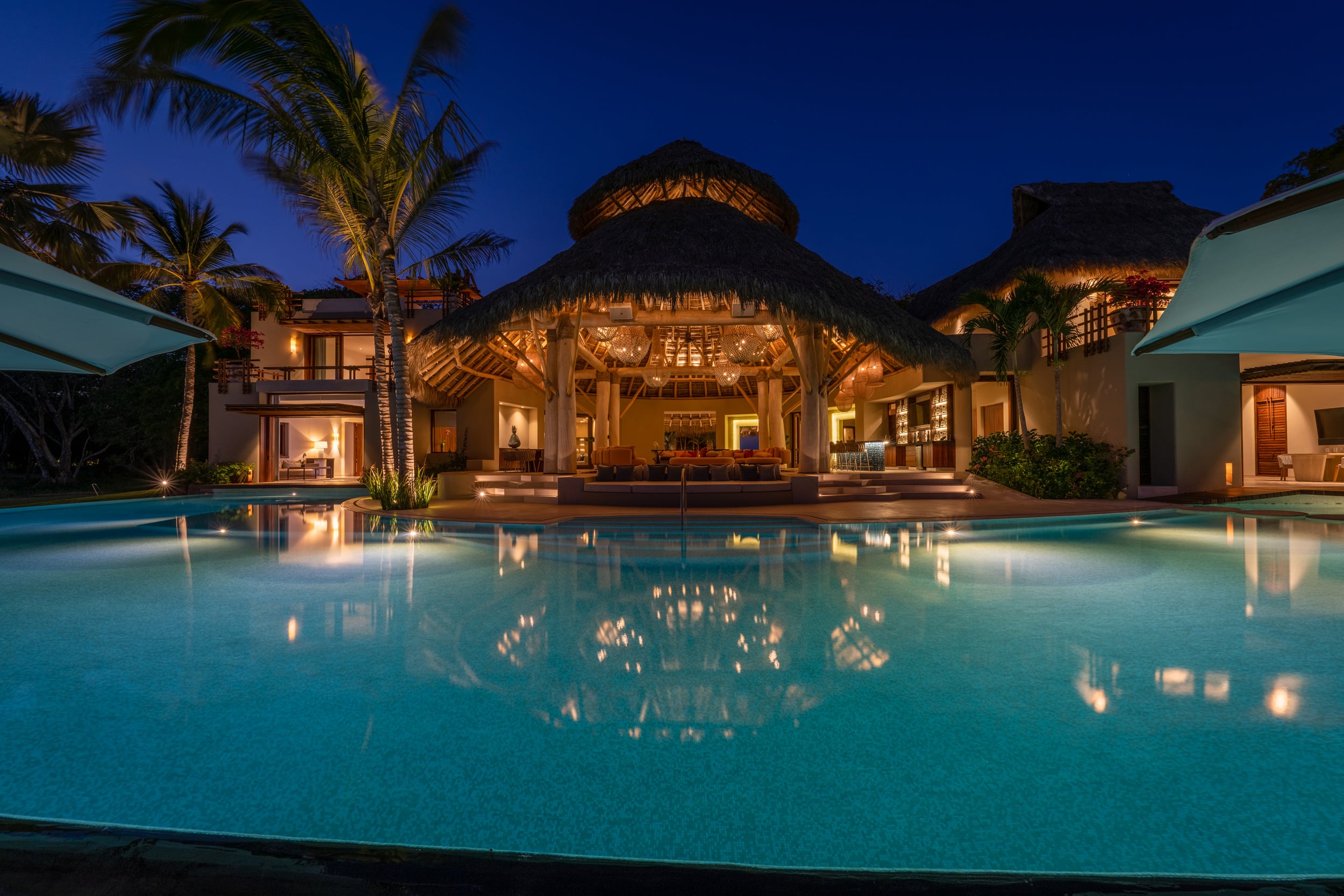 Night view of the main palapa pavilion lit from within, its reflection glowing across the length of the full pool, flanked by illuminated palm trunks and the darkened beach beyond, Casa Akama, Ranchos Estates, Punta Mita.