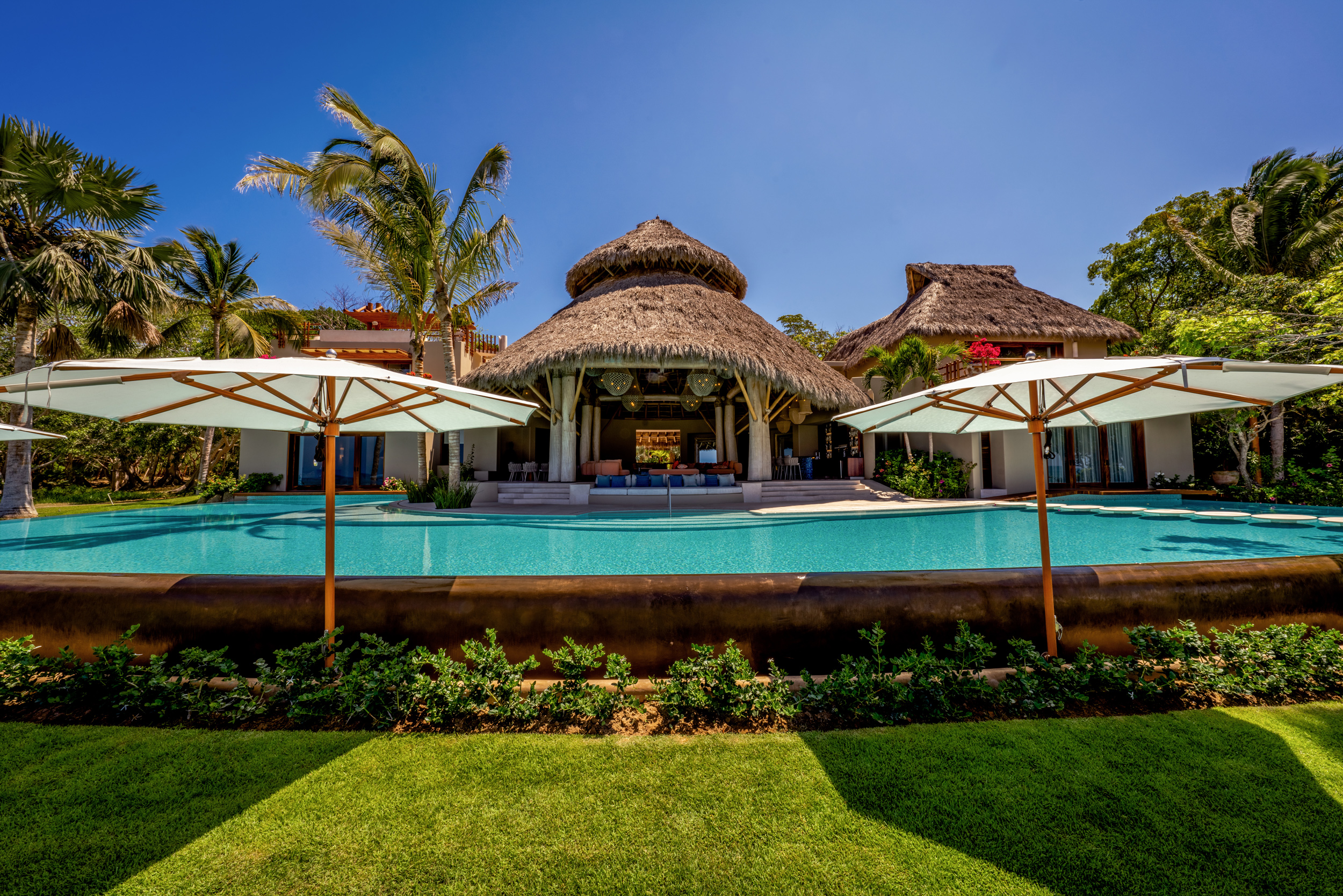 Daytime exterior view of the full main pool with white umbrellas, manicured lawn, the palapa pavilion at center, and surrounding guest wing structures under a deep blue sky, Casa Akama, Ranchos Estates, Punta Mita.