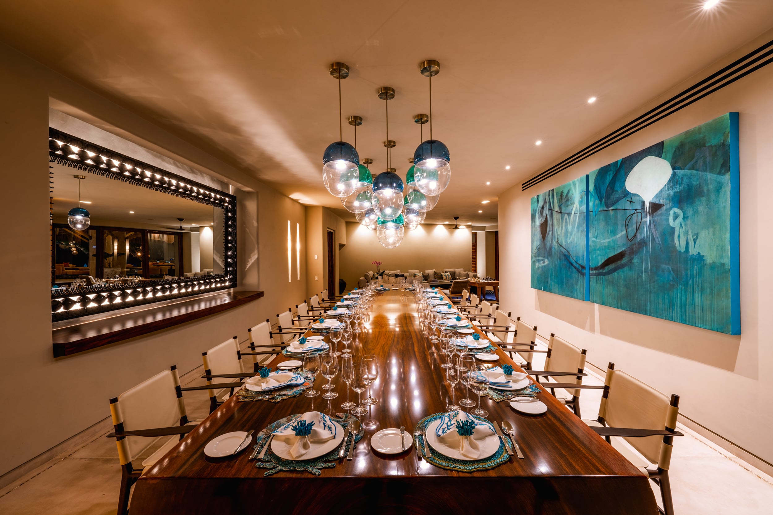 Long formal dining table set for twenty guests with crystal stemware, white china, and teal napkin accents beneath a cluster of glass globe pendants, large contemporary triptych artwork on the right wall, and a wine service window on the left, Casa Akama, Ranchos Estates, Punta Mita.