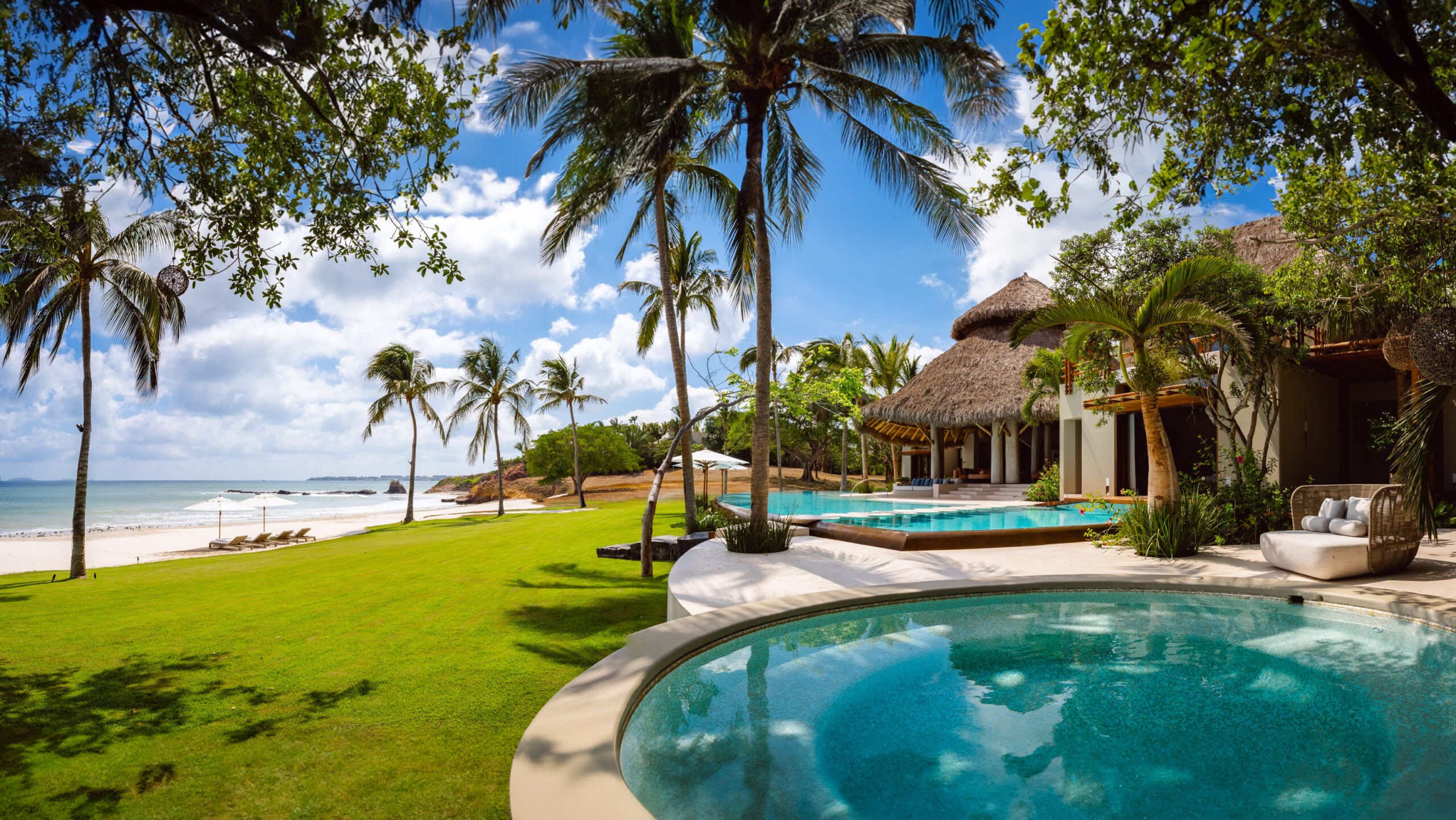 Aerial-angle view from a shaded garden terrace showing the circular plunge pool in the foreground, main infinity pool mid-ground, beachfront lawn, palapa structures, and the Pacific Ocean meeting the Punta Mita coastline, Casa Akama, Ranchos Estates, Punta Mita.