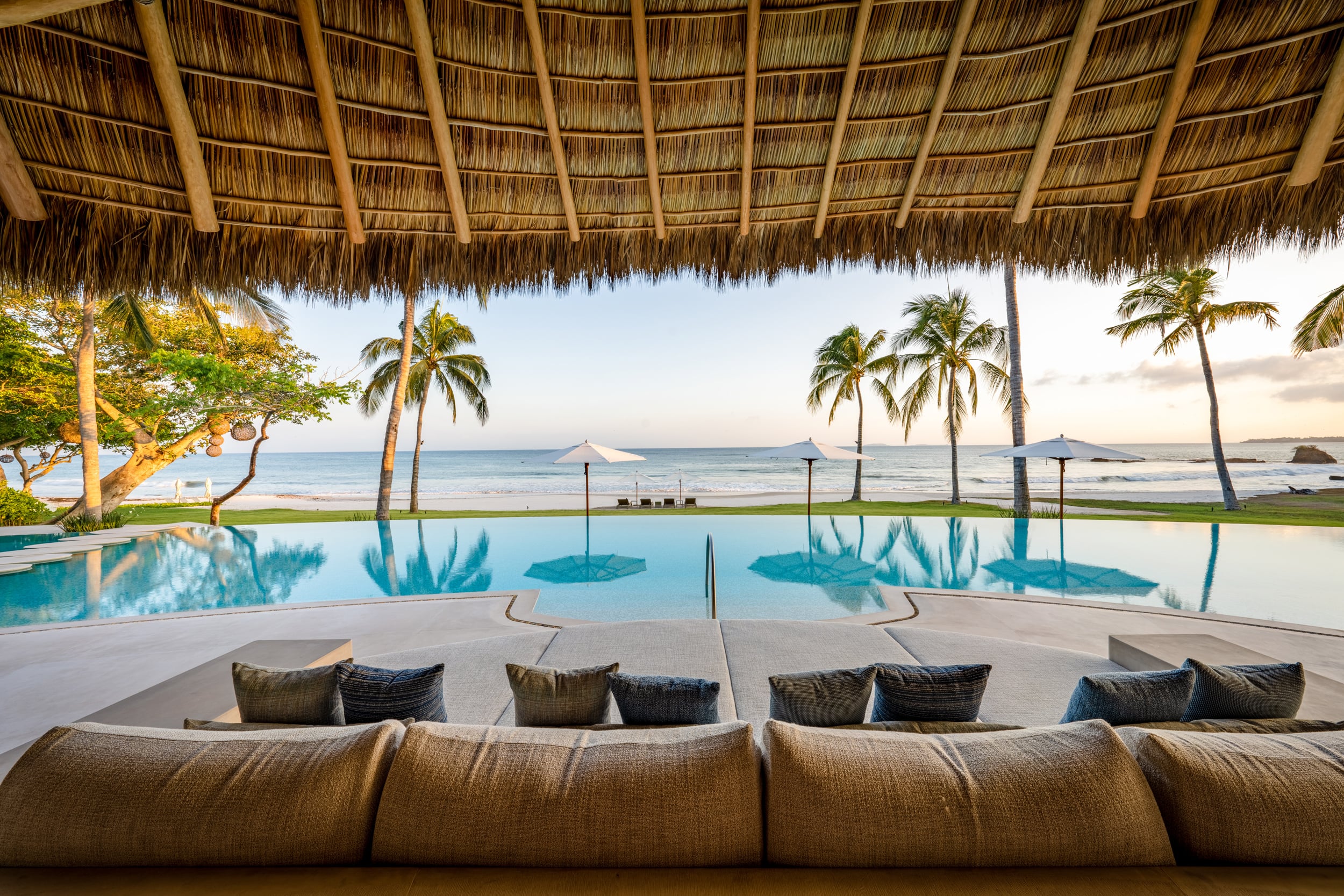 View from a built-in palapa-shaded sofa looking across the still infinity pool toward three beach umbrellas and the Pacific Ocean at early evening with warm golden light on the horizon, Casa Akama, Ranchos Estates, Punta Mita.