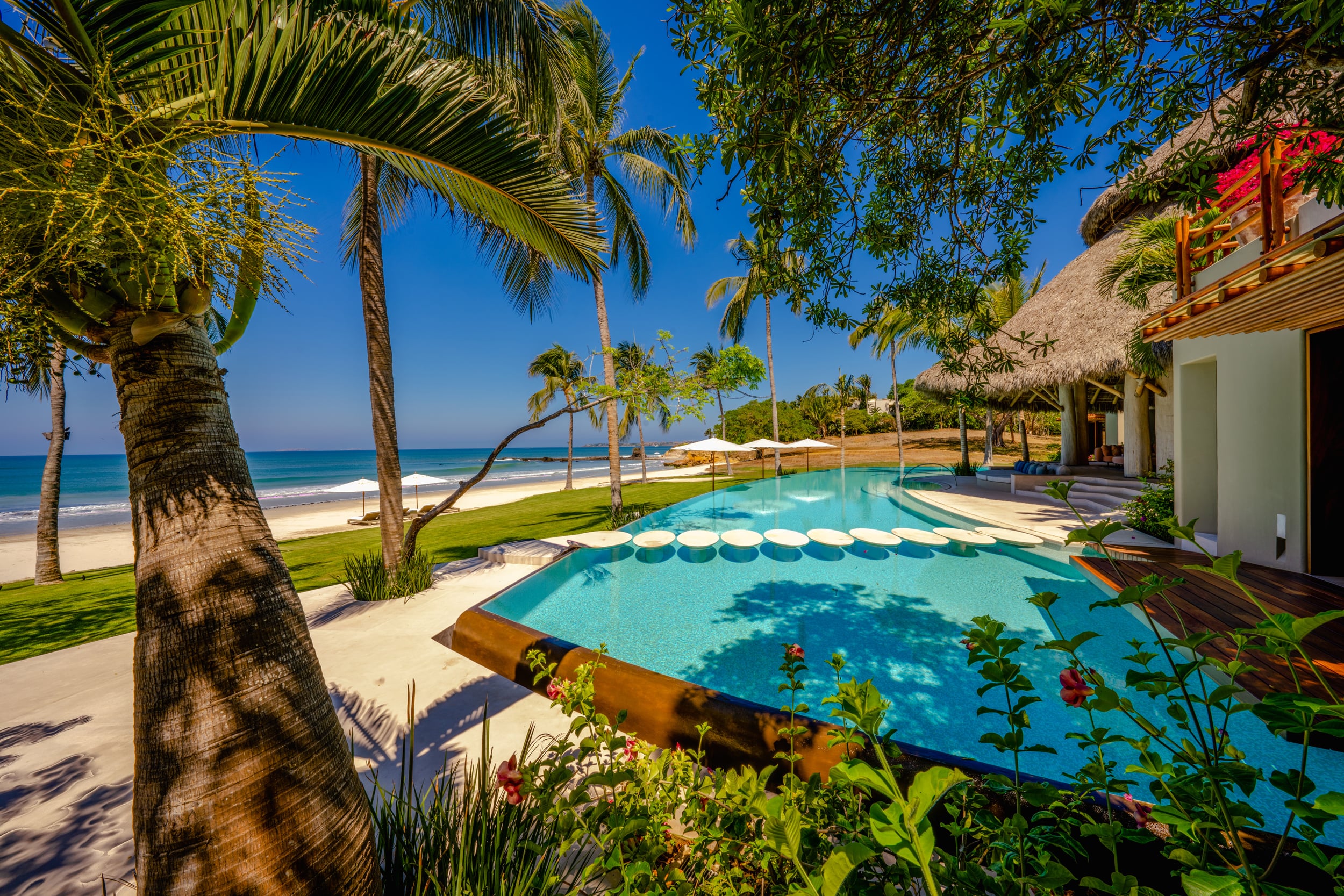 Pool terrace framed by a leaning palm trunk in the foreground, the main infinity pool with a row of floating oval stepping stones along the far edge, and the Pacific beach visible beyond tropical plantings, Casa Akama, Ranchos Estates, Punta Mita.