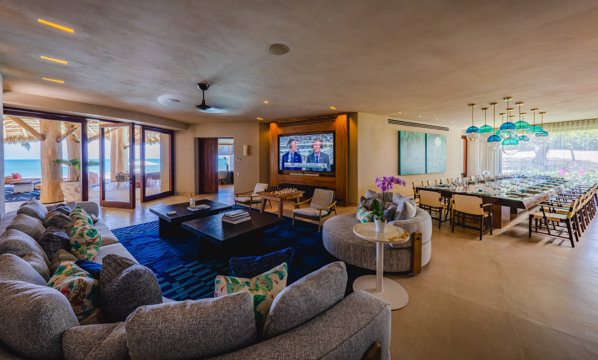 Wide open-plan interior showing a large living area with curved sectional sofas on a blue rug and a wall-mounted television, flowing directly into a full-length formal dining room set for twenty beneath teal pendant lights, with ocean views visible through open doors at the far left, Casa Akama, Ranchos Estates, Punta Mita.