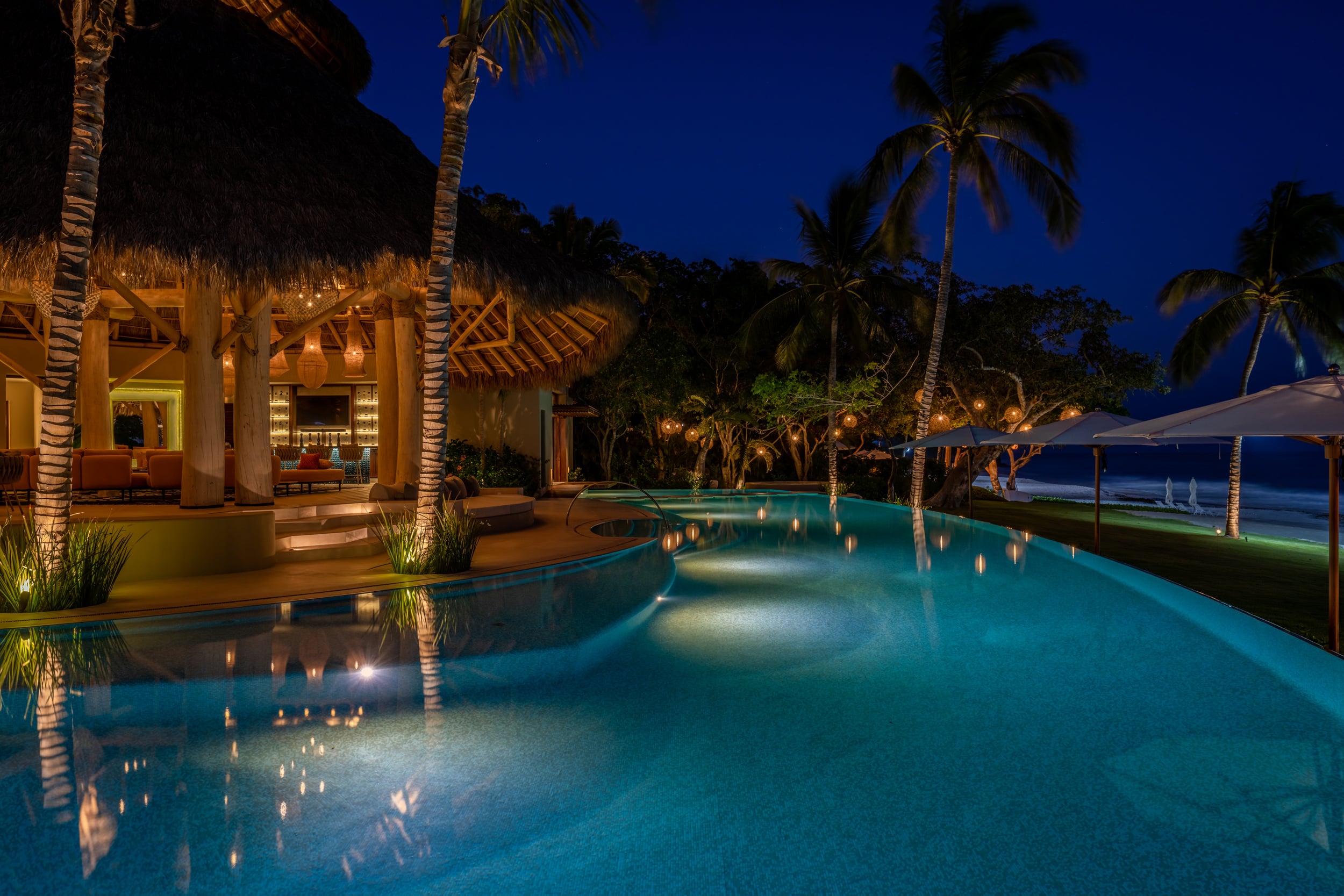 Night view of the main pool lit from below in vivid blue-green tones, with the palapa pavilion glowing amber from interior lighting, candles floating in the water along the edge, and illuminated path lights dotting the garden, Casa Akama, Ranchos Estates, Punta Mita.