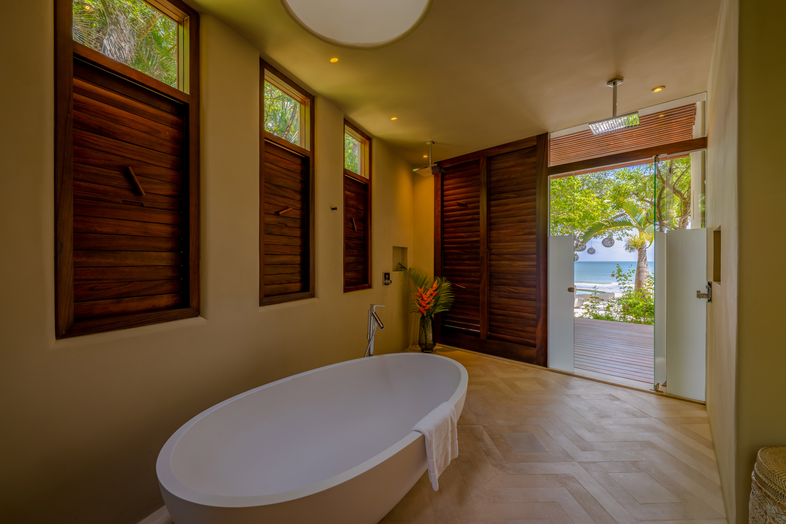 Freestanding oval soaking tub in a spa bathroom with herringbone-tile floor, wood-louvered shutters on three sides letting in filtered light, an open glass door revealing a beach and palm fronds beyond, and a ceiling-mounted rain showerhead above, Casa Akama, Ranchos Estates, Punta Mita.